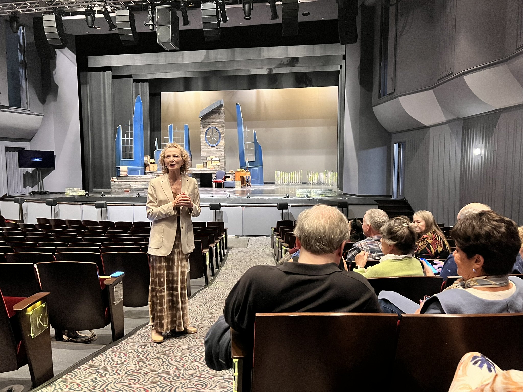 Female standing in theater speaking to a group of others in chairs.
