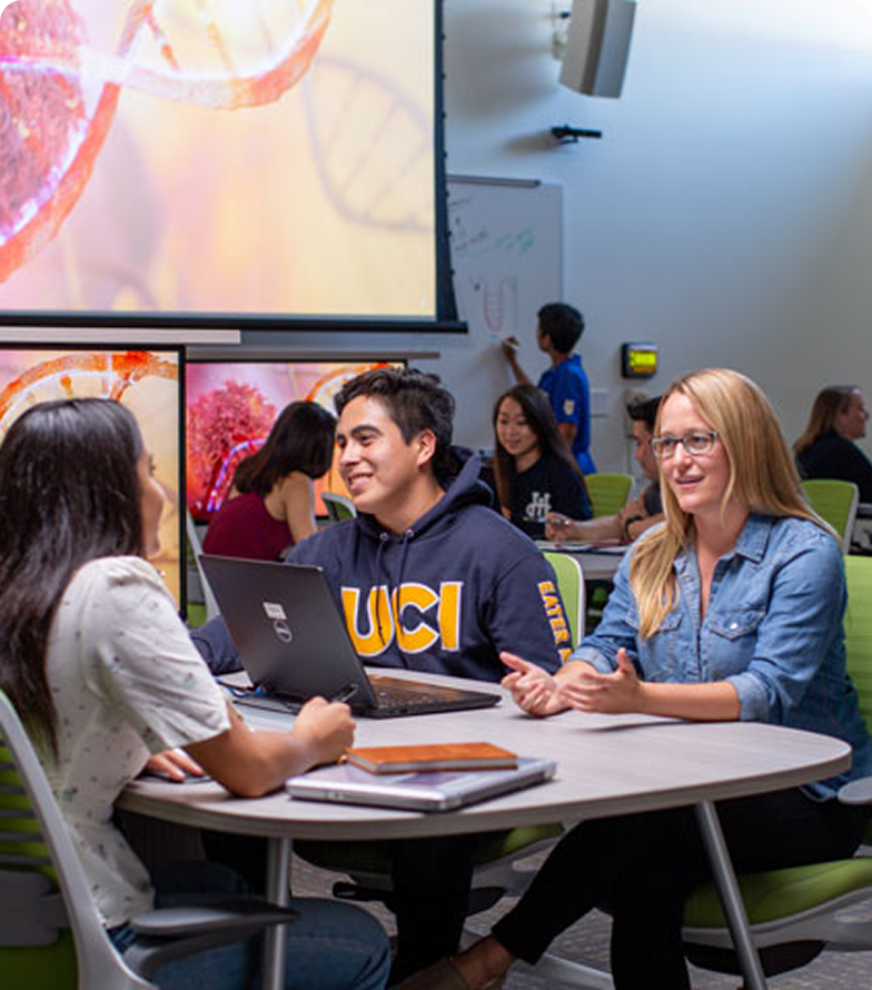 “Three students seated at a round table in a modern classroom collaborate with a laptop displaying a ‘UCI’ logo, while others study and write on a whiteboard in the background.”