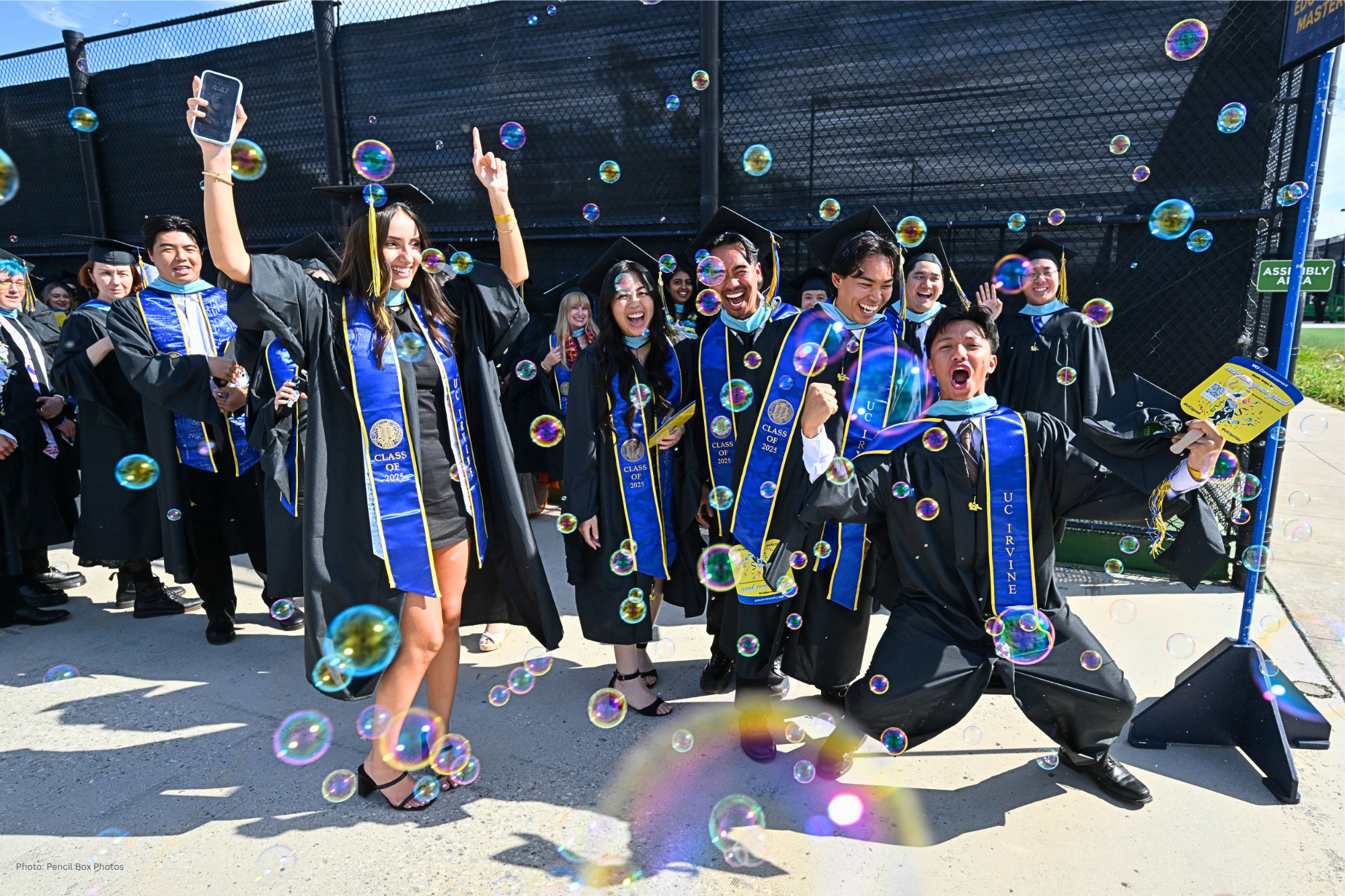 Group of graduates in caps, gowns, and blue stoles posing and celebrating outdoors as bubbles fill the air.