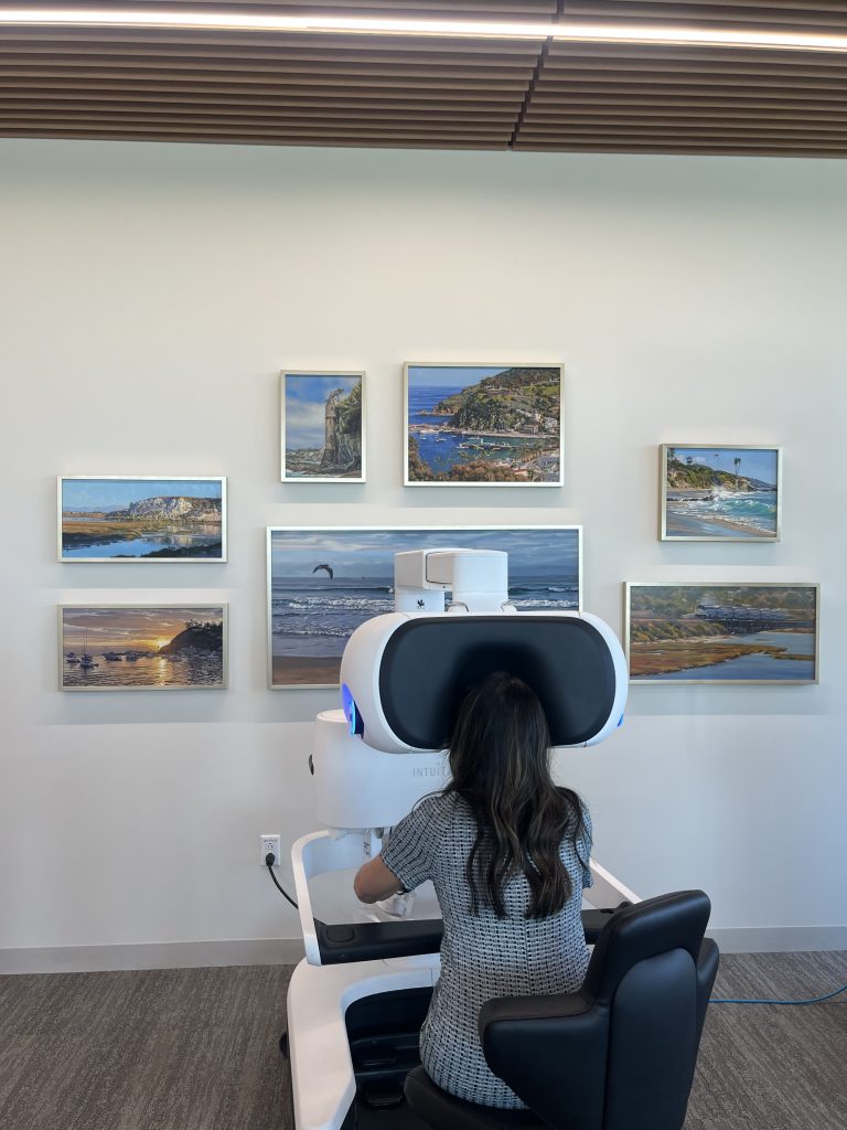 A guest at a hospital places her head in a machine to receive a demonstration of a robotic surgery machine. A collage of artwork shows pictures of California coasts on the wall in the background.