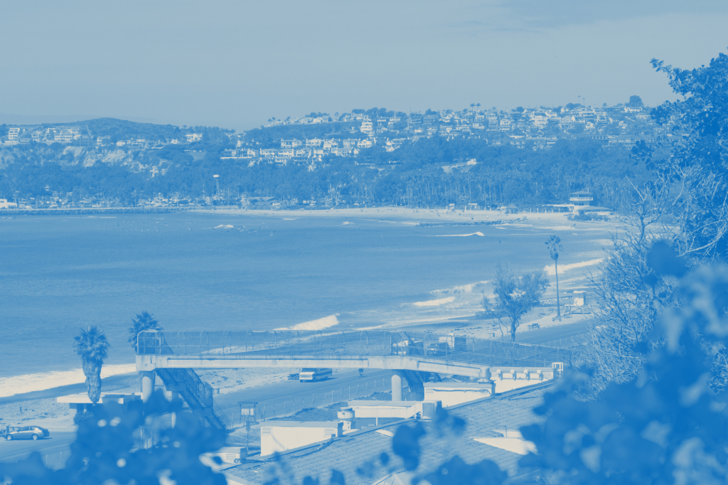 A panoramic view of a coastal town with buildings on hills overlooking a beach and ocean with waves breaking on the shore.