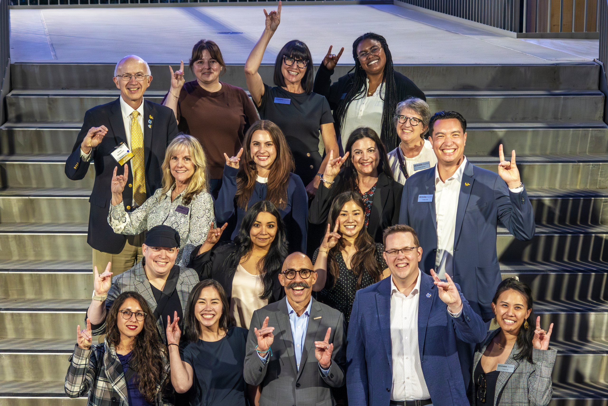 Group of UC Irvine alumni pose together on outdoor steps, wearing name tags and making hand signs at an event.