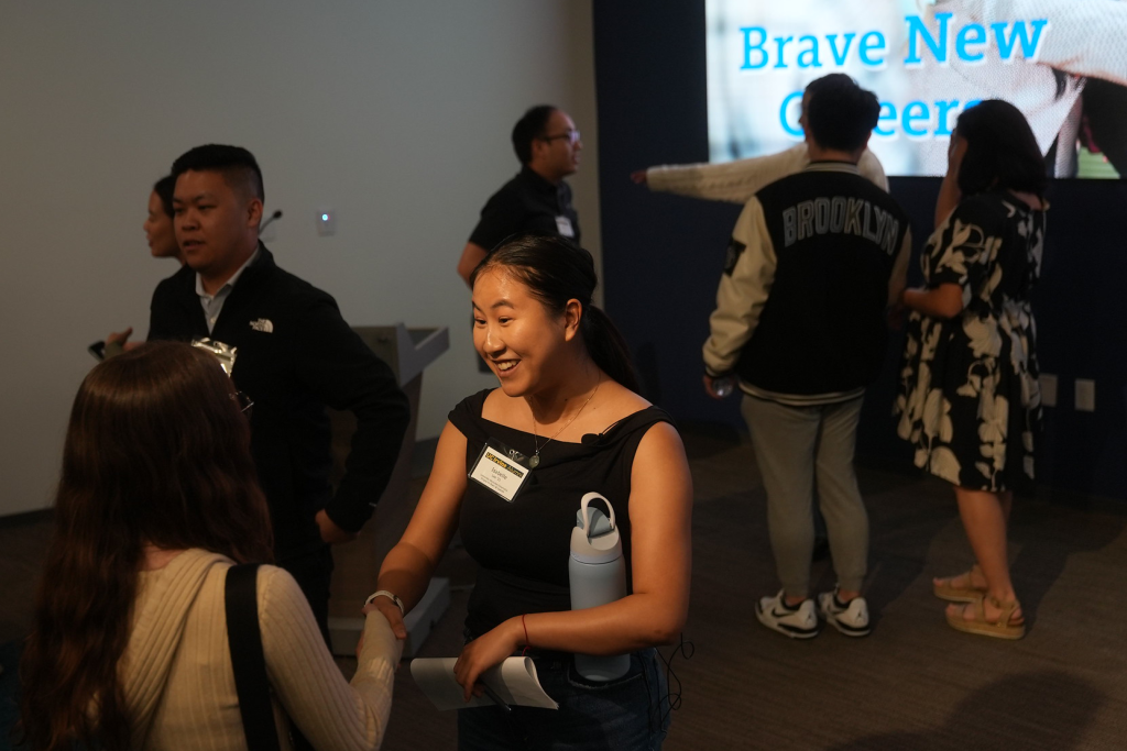 People networking at an indoor career services event. In the foreground, two attendees shake hands and talk, while several others converse in small groups around the room.