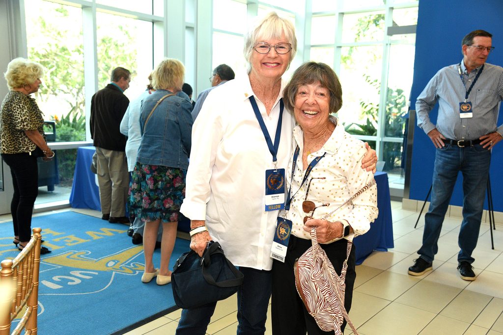 Two event attendees wearing blue lanyards pose together inside a bright reception space with floor-to-ceiling windows, while other guests mingle in the background.