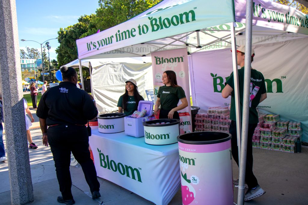 People staff a Bloom booth and hand out beverages under a tent at an outdoor campus event.