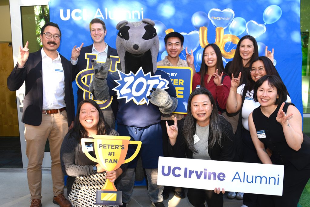 UC Irvine alumni pose with the mascot at an event, holding props and a sign that reads “UC Irvine Alumni.”