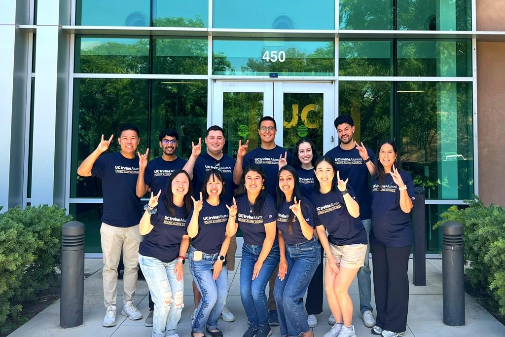 Group of UC Irvine alumni stand outside a campus building wearing matching shirts and making hand signs.