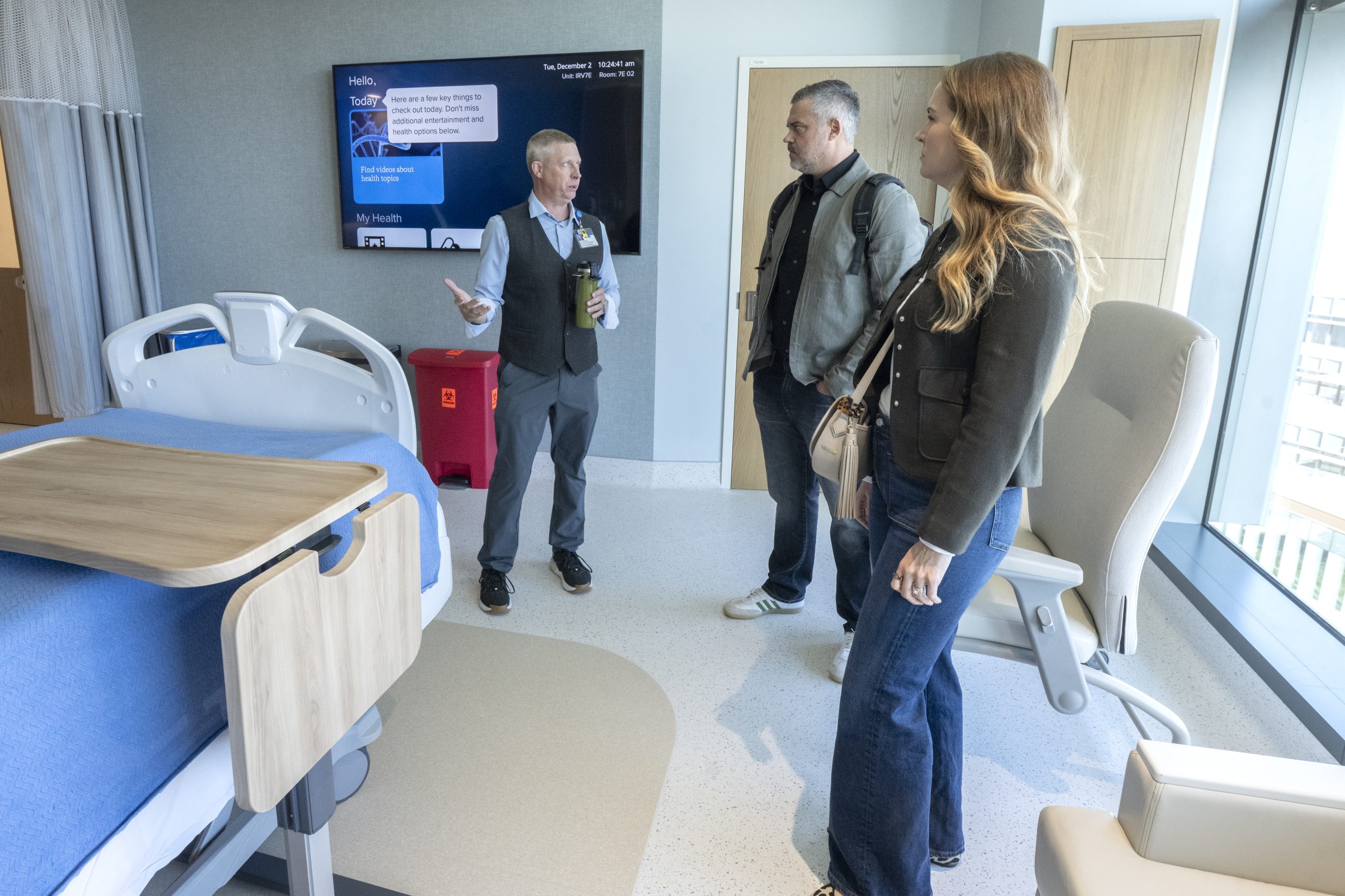A couple listens to a hospital worker talk while standing in a hospital room with a large TV screen on the wall.
