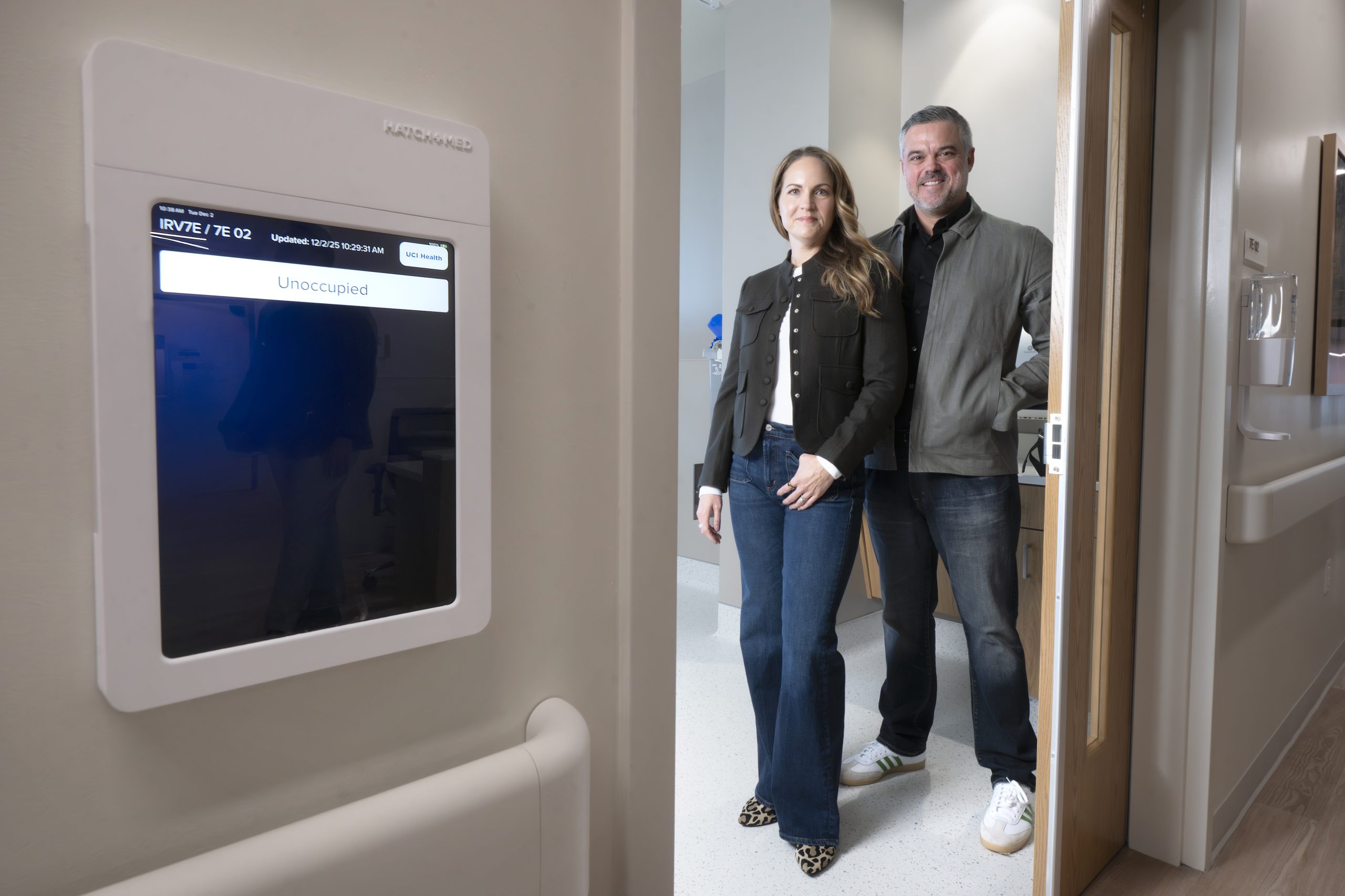 A couple poses in the doorway of a hospital room next to a digital screen