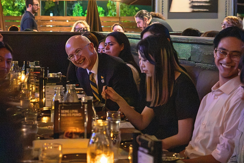 People seated at a long candlelit table in a dim restaurant setting, eating and talking during a group dinner.