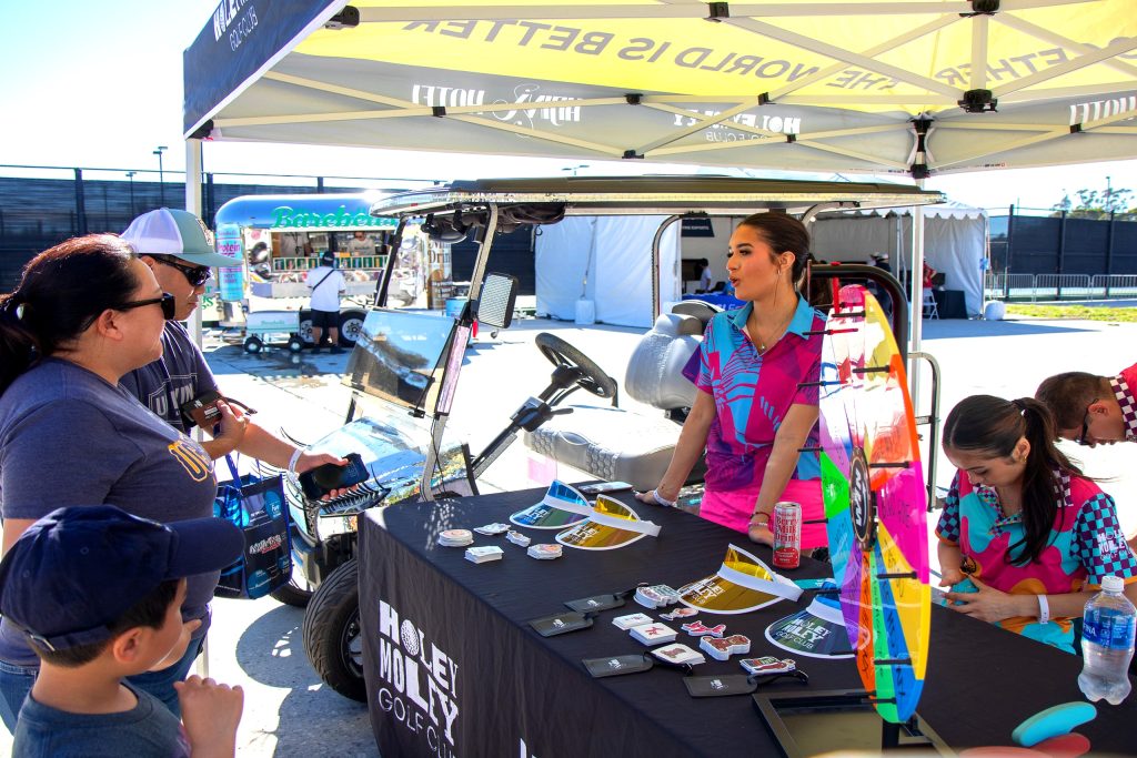 A woman stands behind a booth table under a tent, talking to visitors, with promotional items and a wheel game displayed on the table.