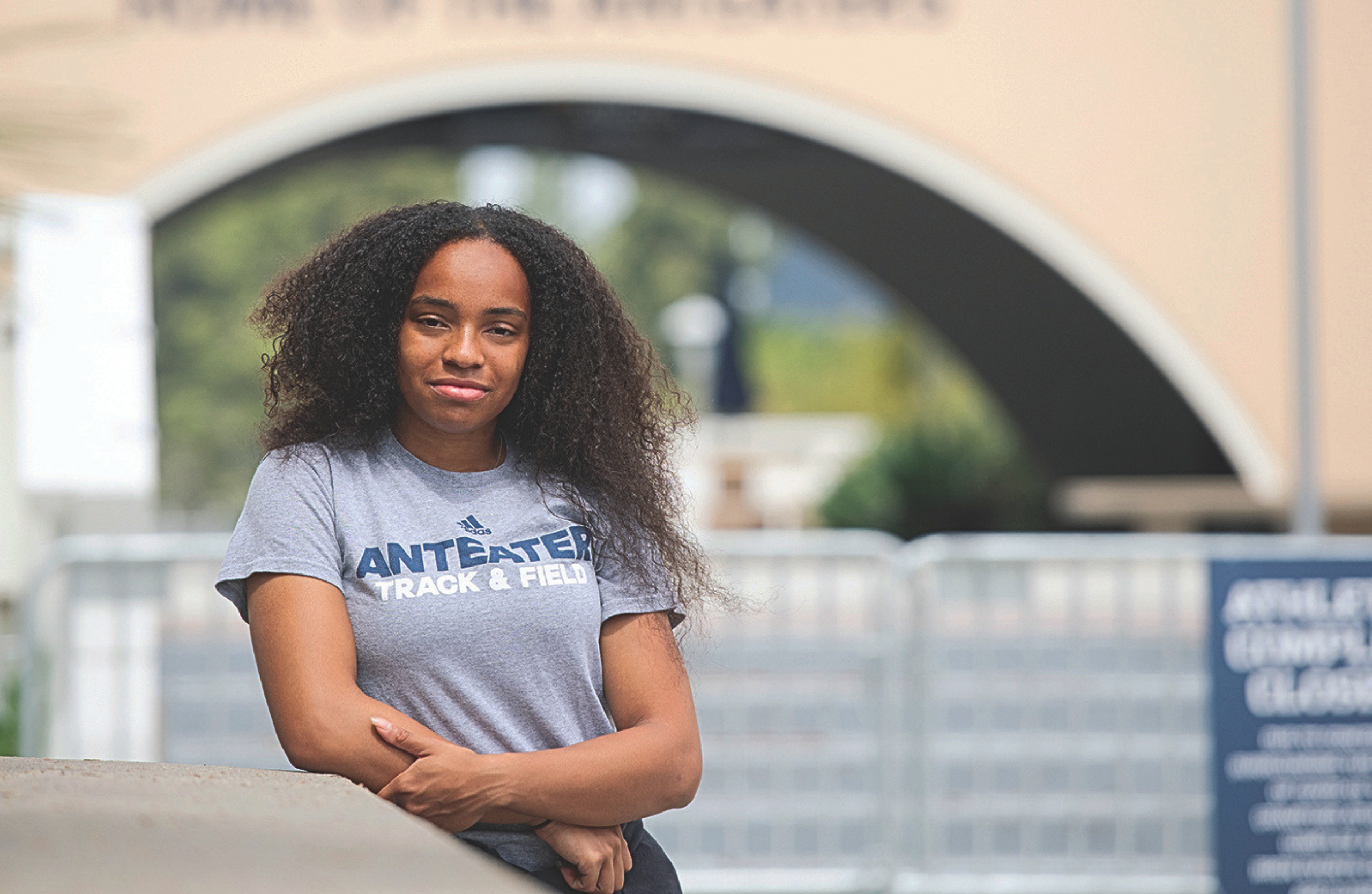 Person wearing an “Anteater Track & Field” T-shirt leaning on a railing in front of a large arched campus walkway.