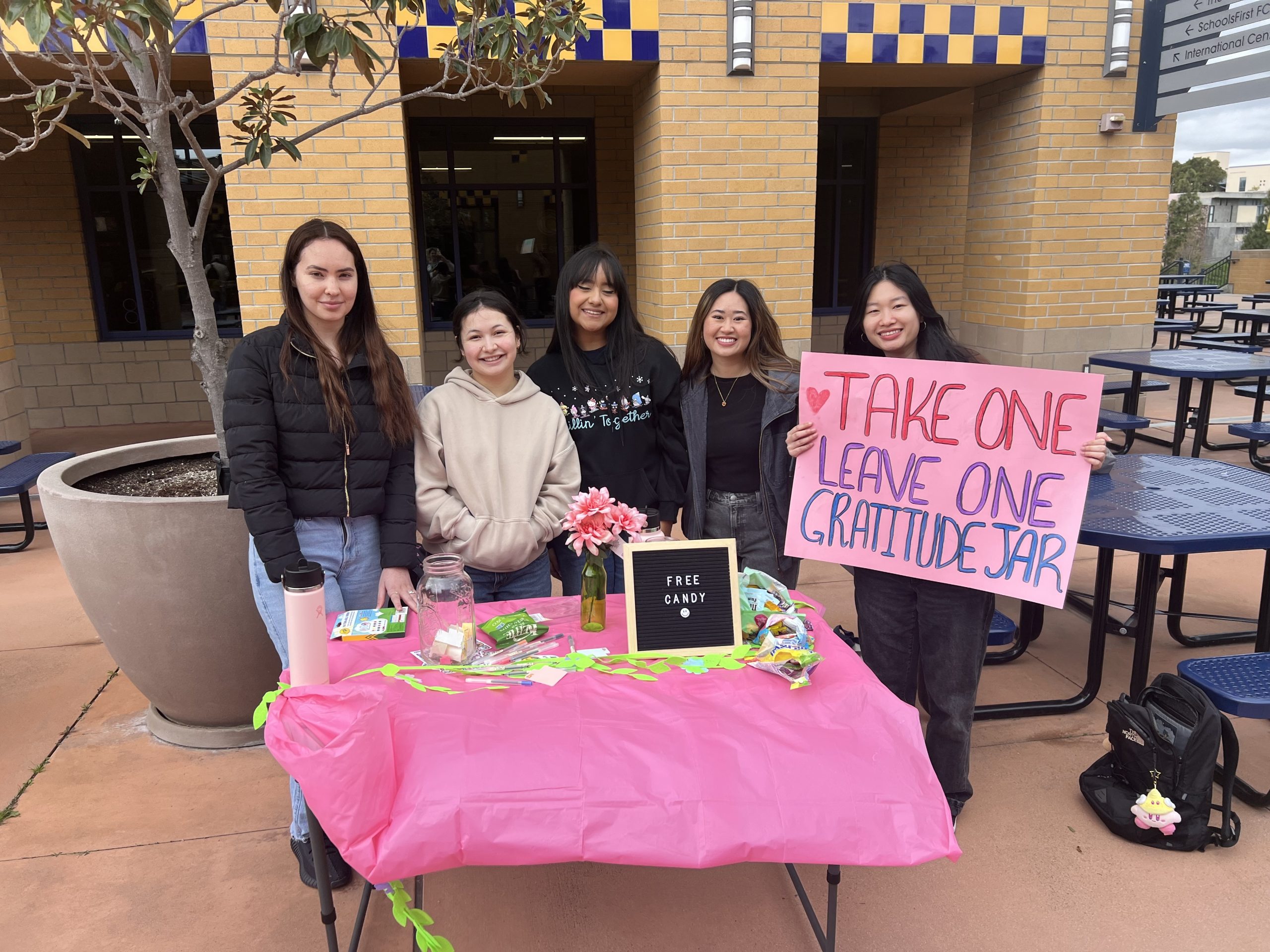 Students pose in front of a table with a pink tablecloth and hold a sign saying "Take One Leave One Gratitude Jar" during Happiness Week