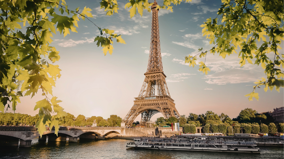 The Eiffel Tower at sunset, framed by green leaves, stands prominently against a pink sky. Below, a river with a passing boat adds calmness.
