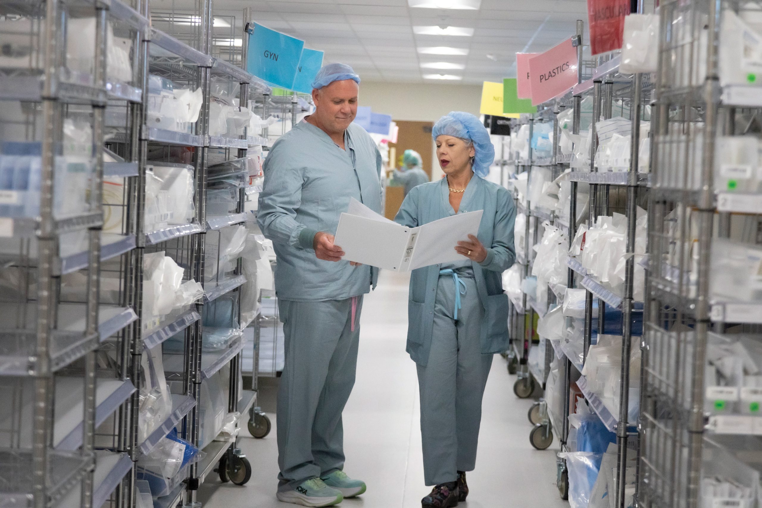 Two hospital workers in scrubs and caps review papers in a binder in the middle of an aisle lined with medical supplies.