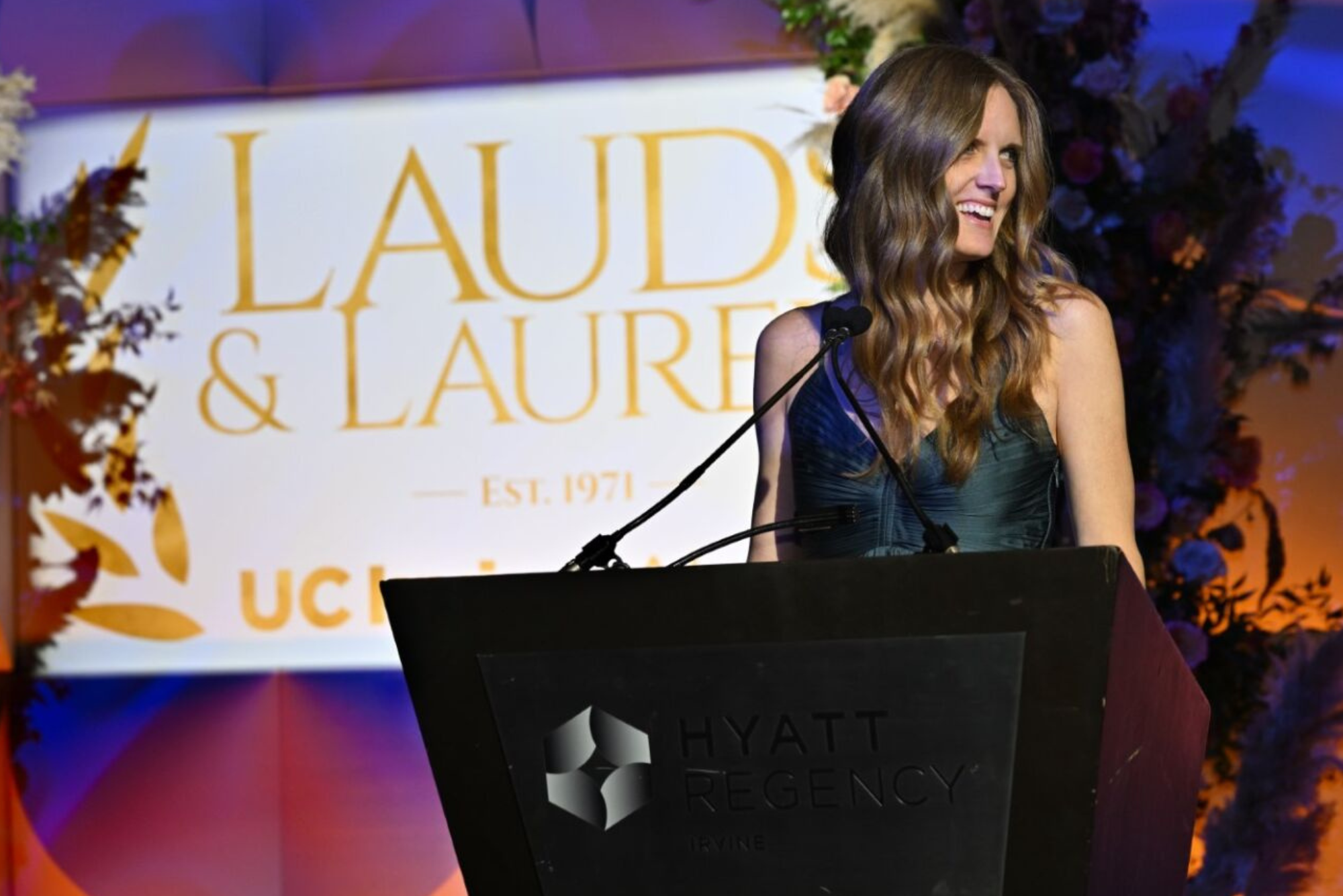 Speaker at a podium during a UC Irvine Alumni Association awards event, standing in front of a "Lauds & Laurels" backdrop.