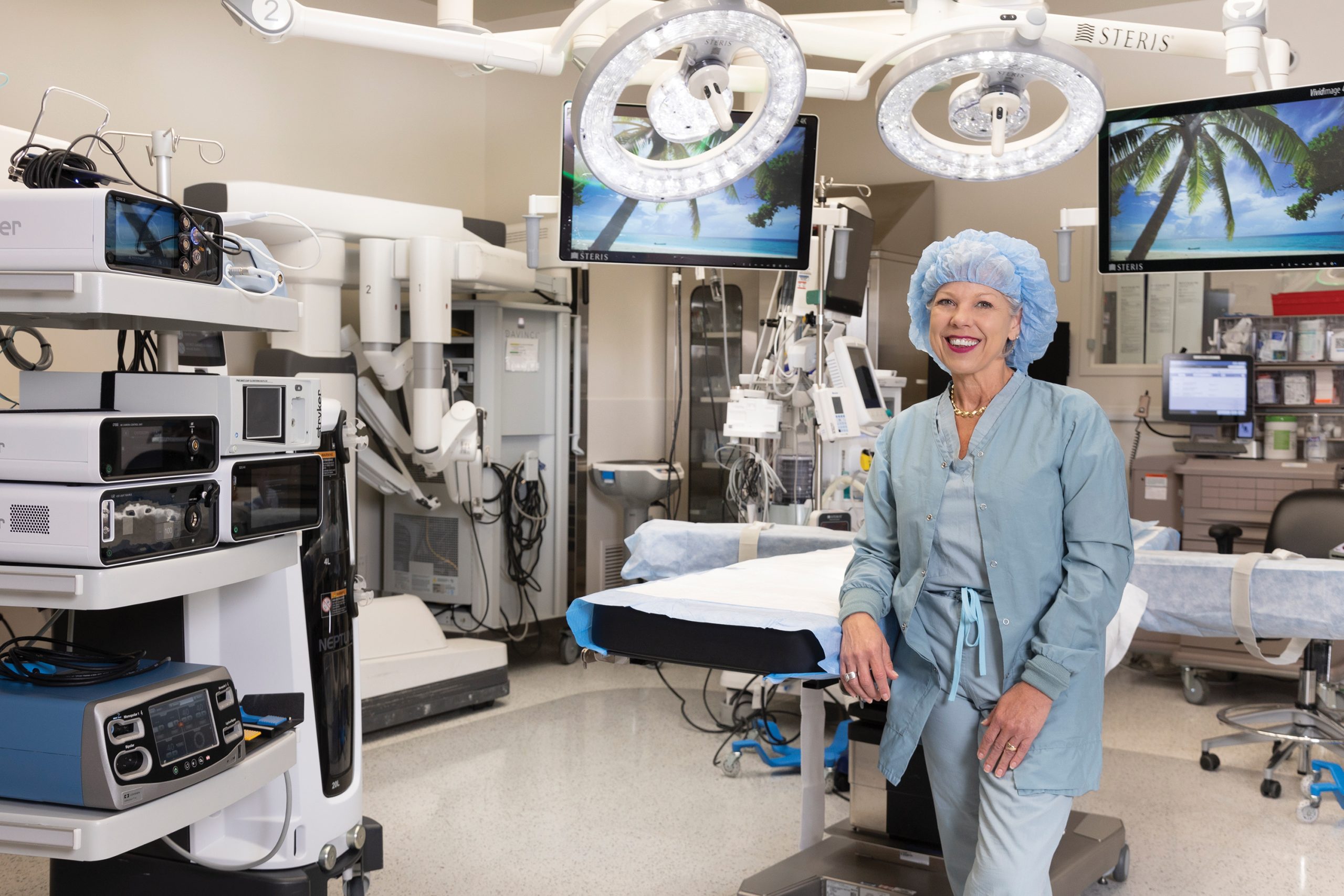 A hospital worker in scrubs and cap poses in an operating room filled with equipment