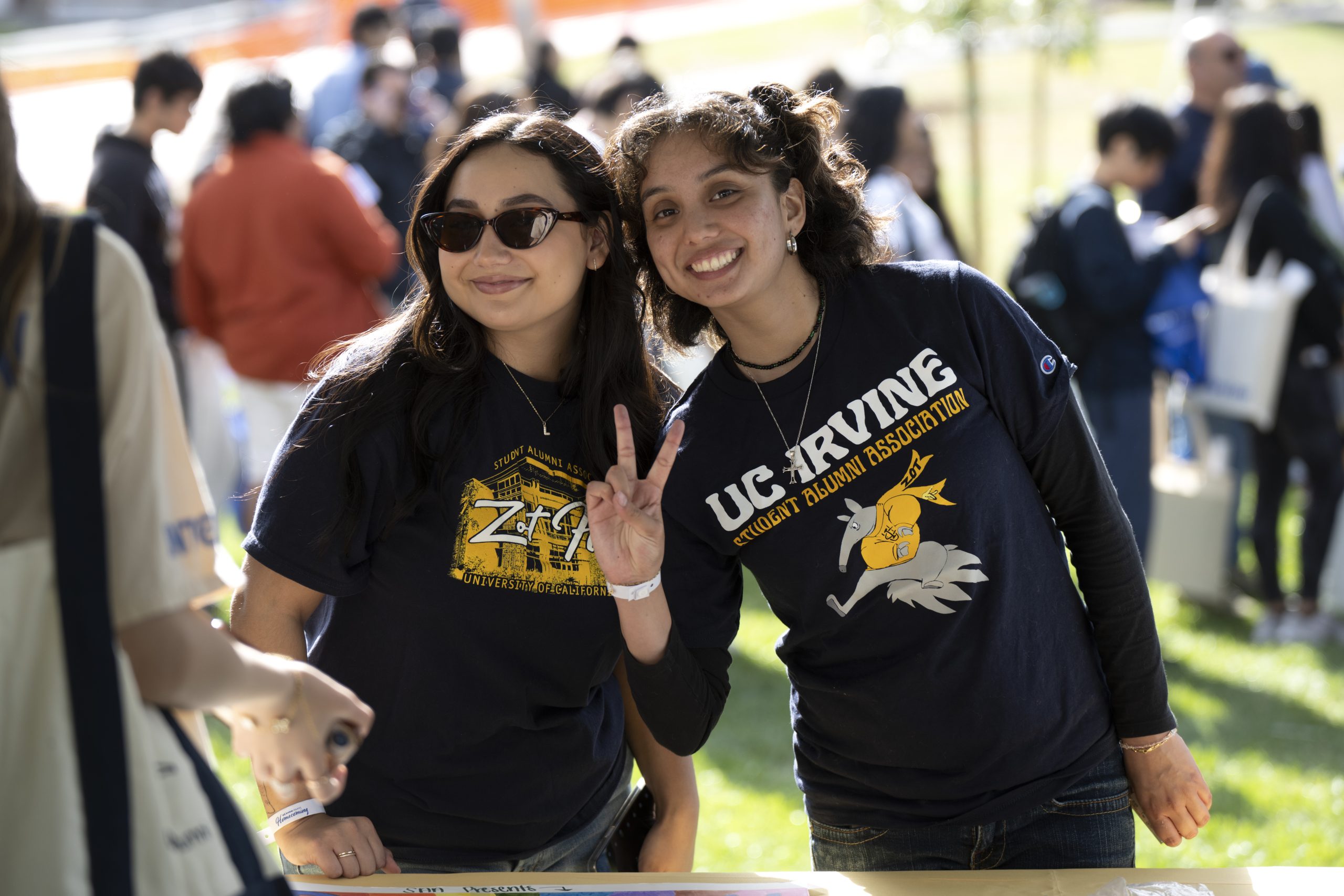 Two people in UC Irvine Student Alumni Association T-shirts stand together at an outdoor campus event booth, with one raising a peace sign and others gathered in the background.