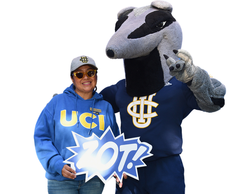 A person wearing a blue UCI hoodie and UC Irvine cap stands next to the UC Irvine Anteater mascot while holding a sign that reads “ZOT!”