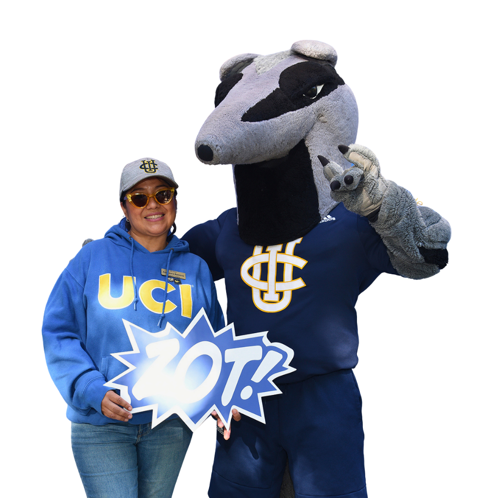 A person wearing a blue UCI hoodie and UC Irvine cap stands next to the UC Irvine Anteater mascot while holding a sign that reads “ZOT!”