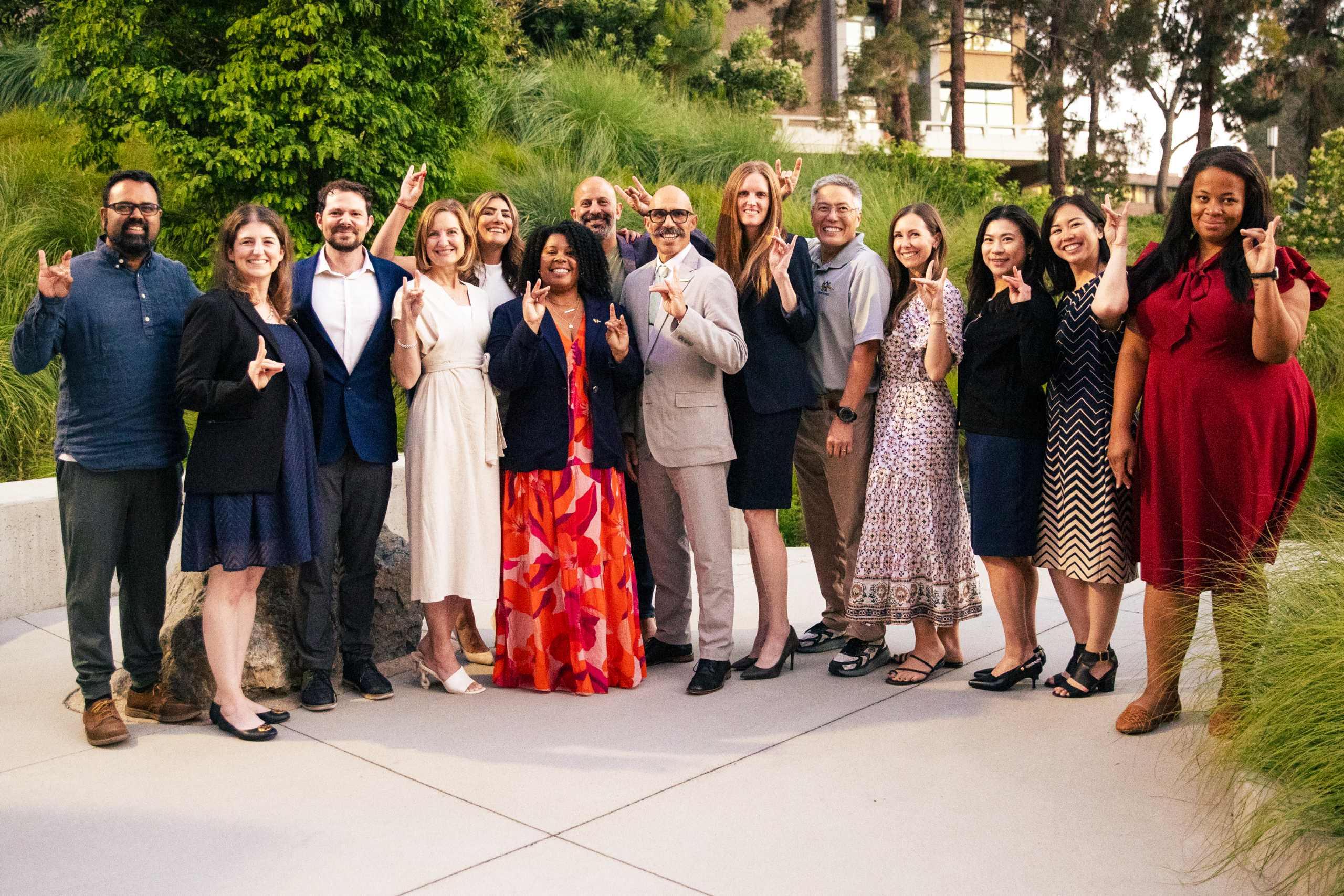 Group portrait of event attendees standing outdoors in a landscaped courtyard, posing together and making hand signs for the camera.