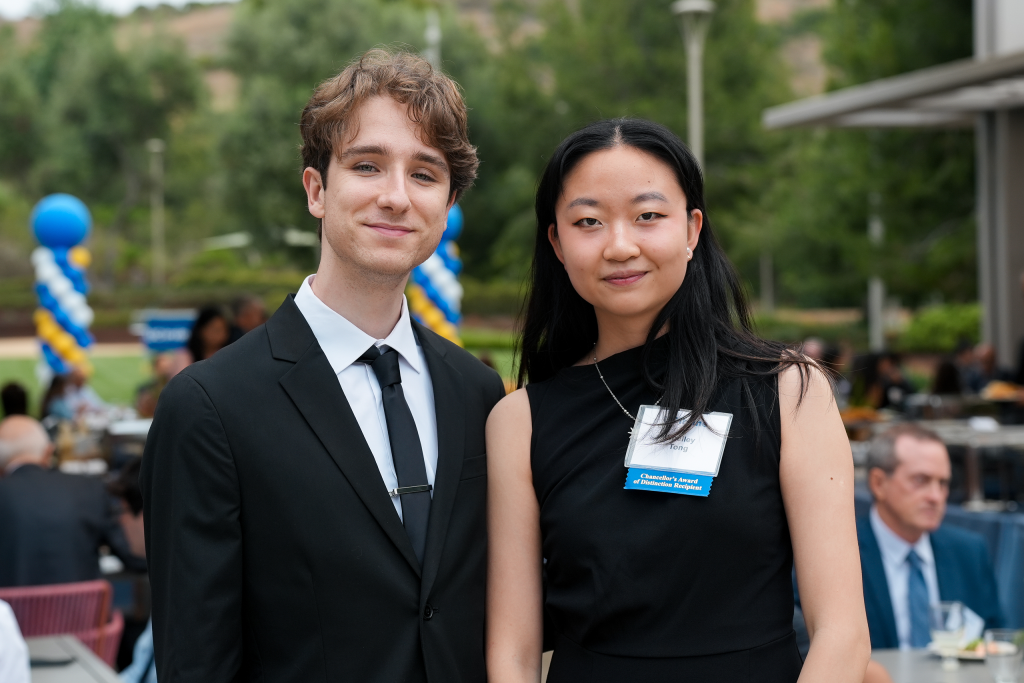Two attendees pose for a portrait at an outdoor recognition event, with tables, guests, and blue-and-gold balloon decorations in the background.