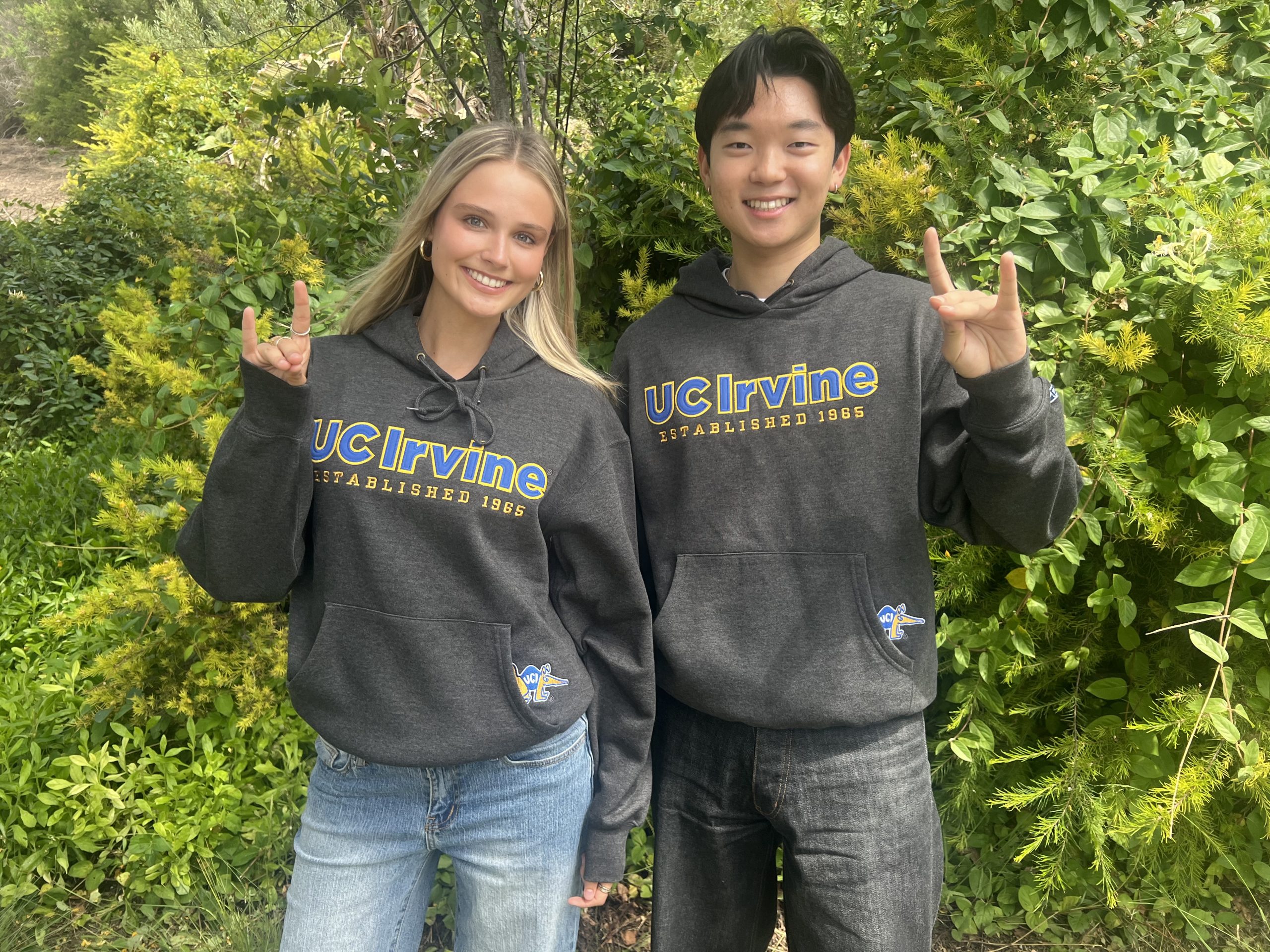 Two people standing outdoors in front of dense green foliage, wearing dark gray UC Irvine hooded sweatshirts with blue and gold lettering reading “UC Irvine Established 1965,” and making hand gestures while facing the camera.