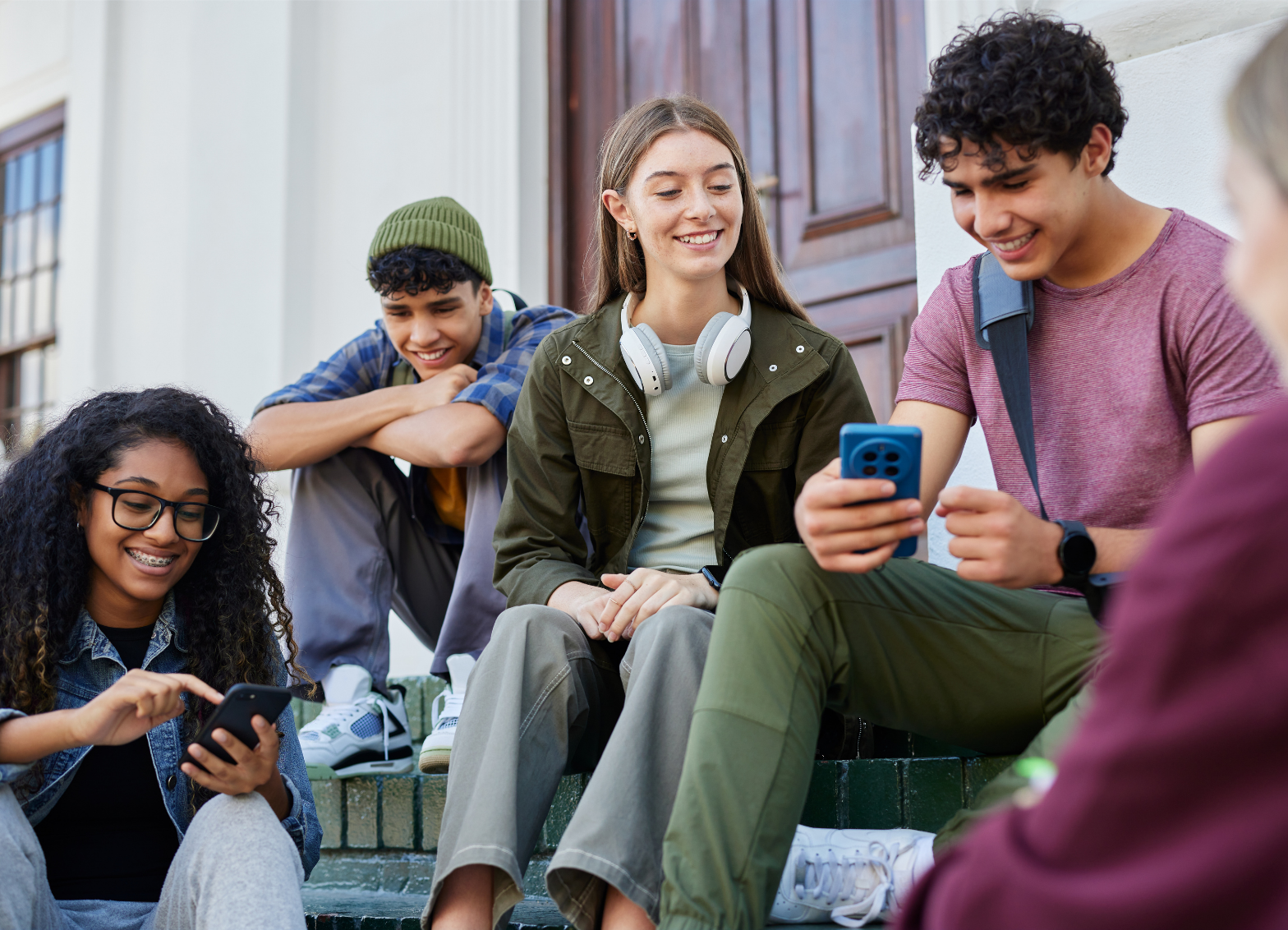 A group of teenagers sits on outdoor steps using smartphones and other mobile devices while gathered together outside a building.