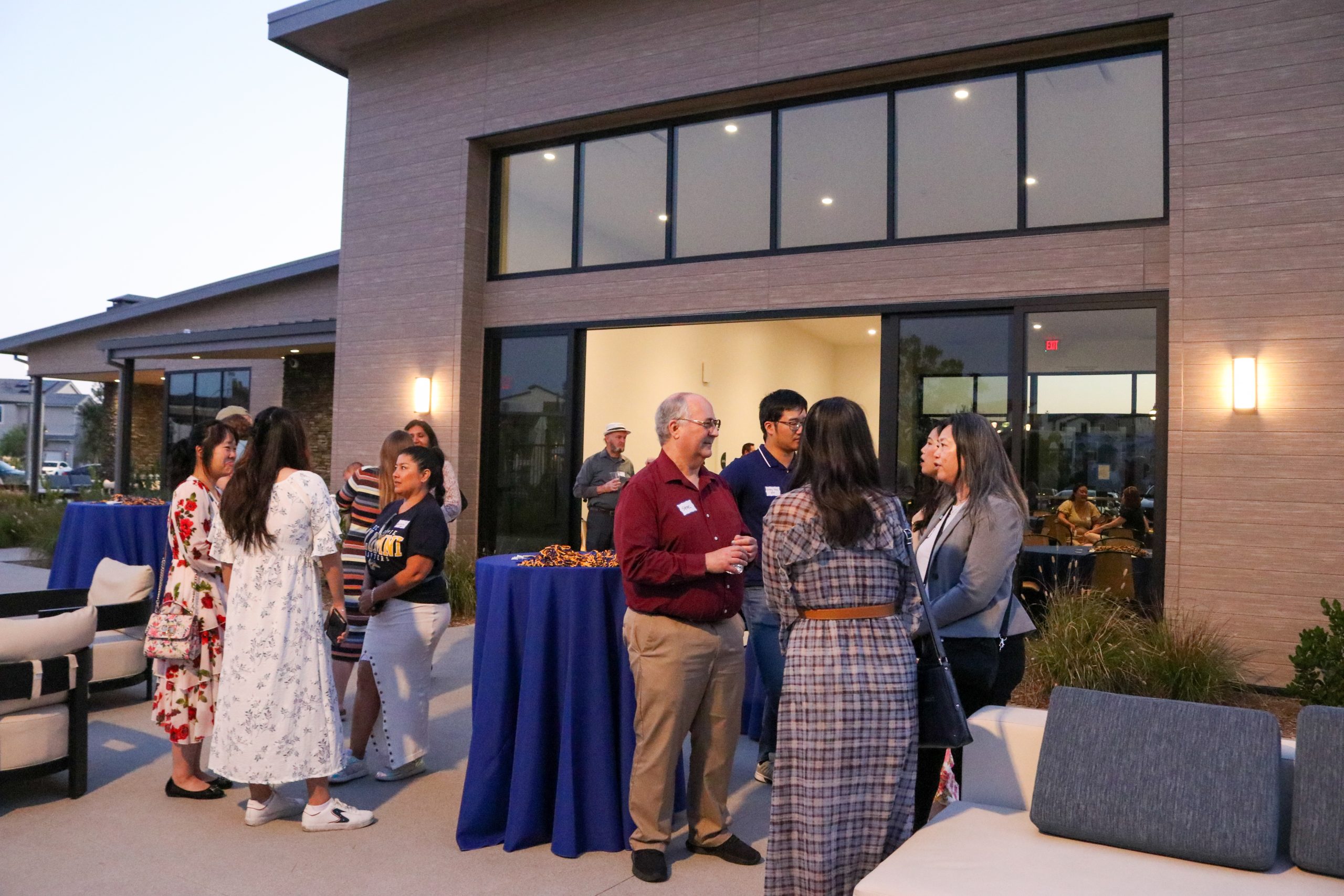 People standing and conversing around cocktail tables on an outdoor patio in front of a modern clubhouse building during an evening networking reception.
