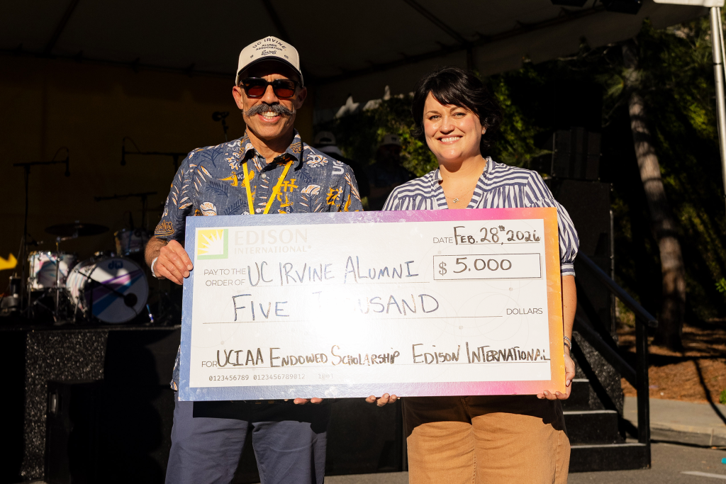 Two event attendees hold a ceremonial $5,000 scholarship donation check for UC Irvine Alumni while standing on an outdoor stage.