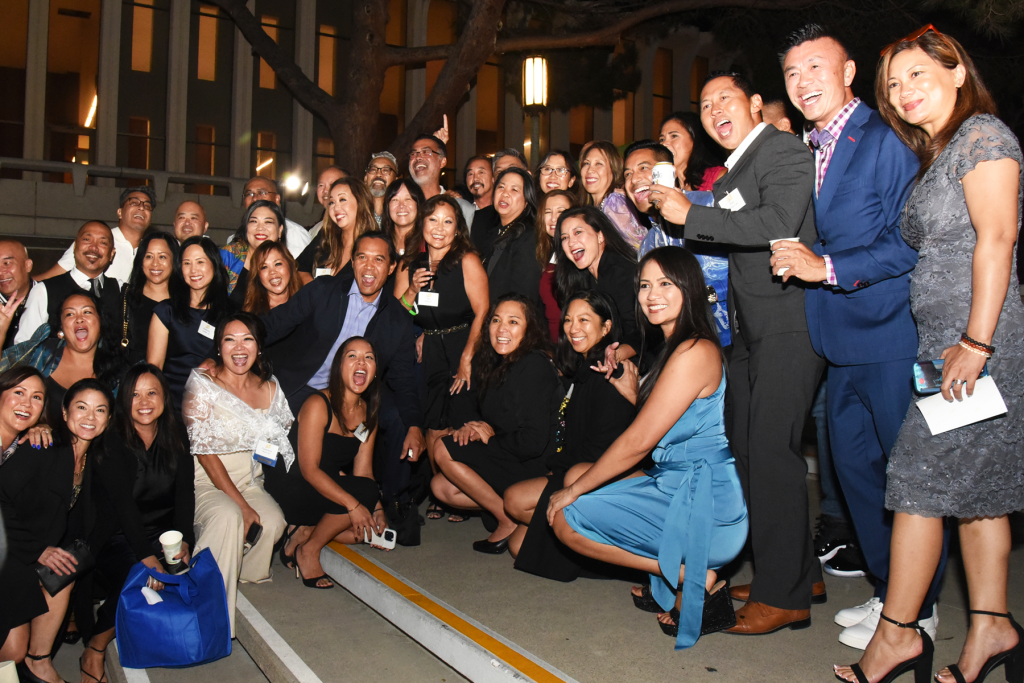Large group of alumni and attendees pose together for a photo at an evening outdoor event, gathered on steps outside a campus building.