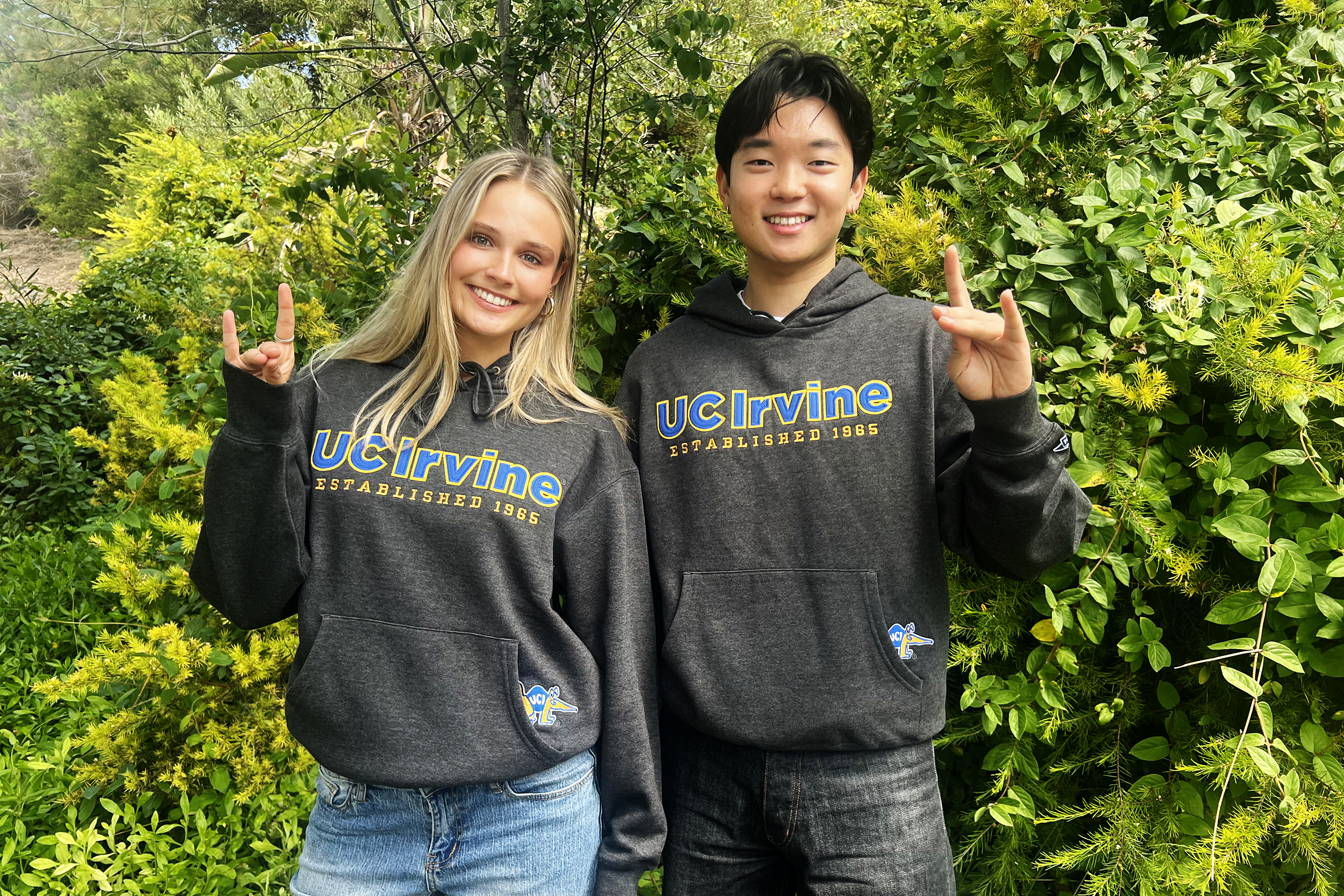 Two people standing outdoors in front of dense green foliage, wearing dark gray UC Irvine hooded sweatshirts with the text “UC Irvine Established 1965” and making hand gestures toward the camera.