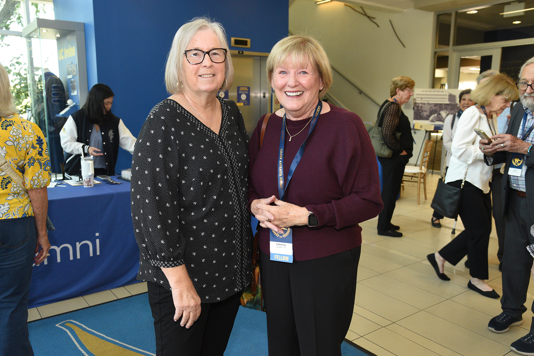 Two attendees pose for a photo at a UC Irvine Alumni Golden Anteater Society event while guests mingle in the background.