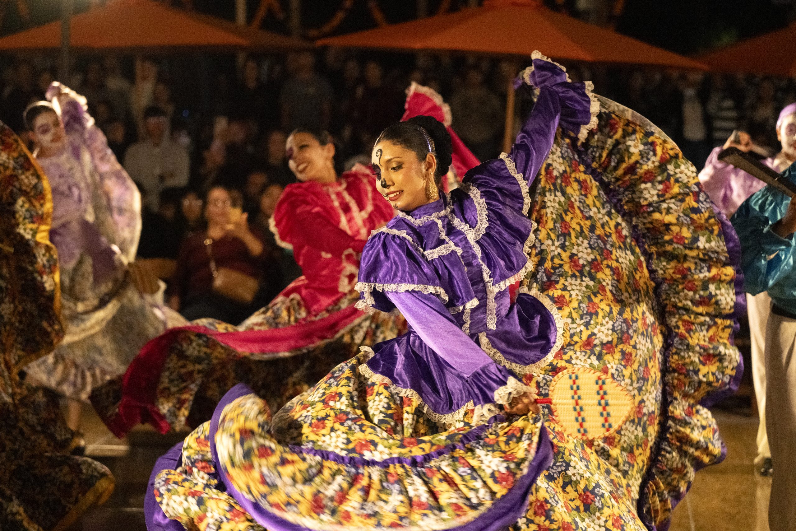Performers in colorful traditional dresses swirl patterned skirts during a cultural dance performance before an audience at an outdoor evening event.
