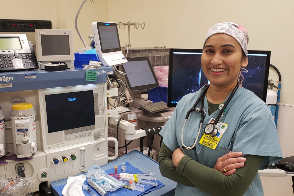 Person in medical scrubs with a stethoscope stands beside anesthesia and patient-monitoring equipment in a hospital procedure room.