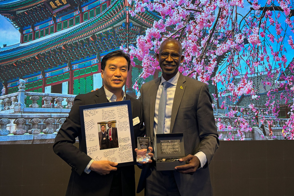 Two individuals in business attire hold recognition awards while standing in front of a large display showing traditional architecture and pink flowering trees.