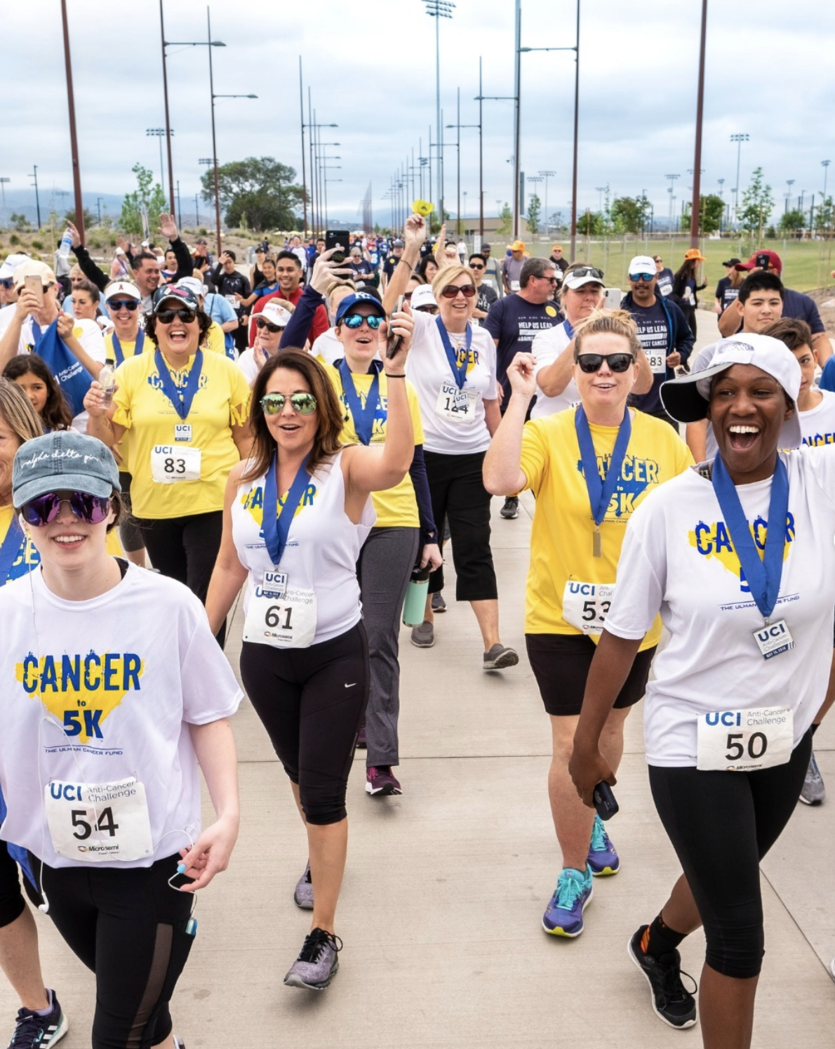 Participants in a UC Irvine Anti-Cancer Challenge 5K walk move along a paved pathway wearing event shirts, race bibs, and finisher medals.