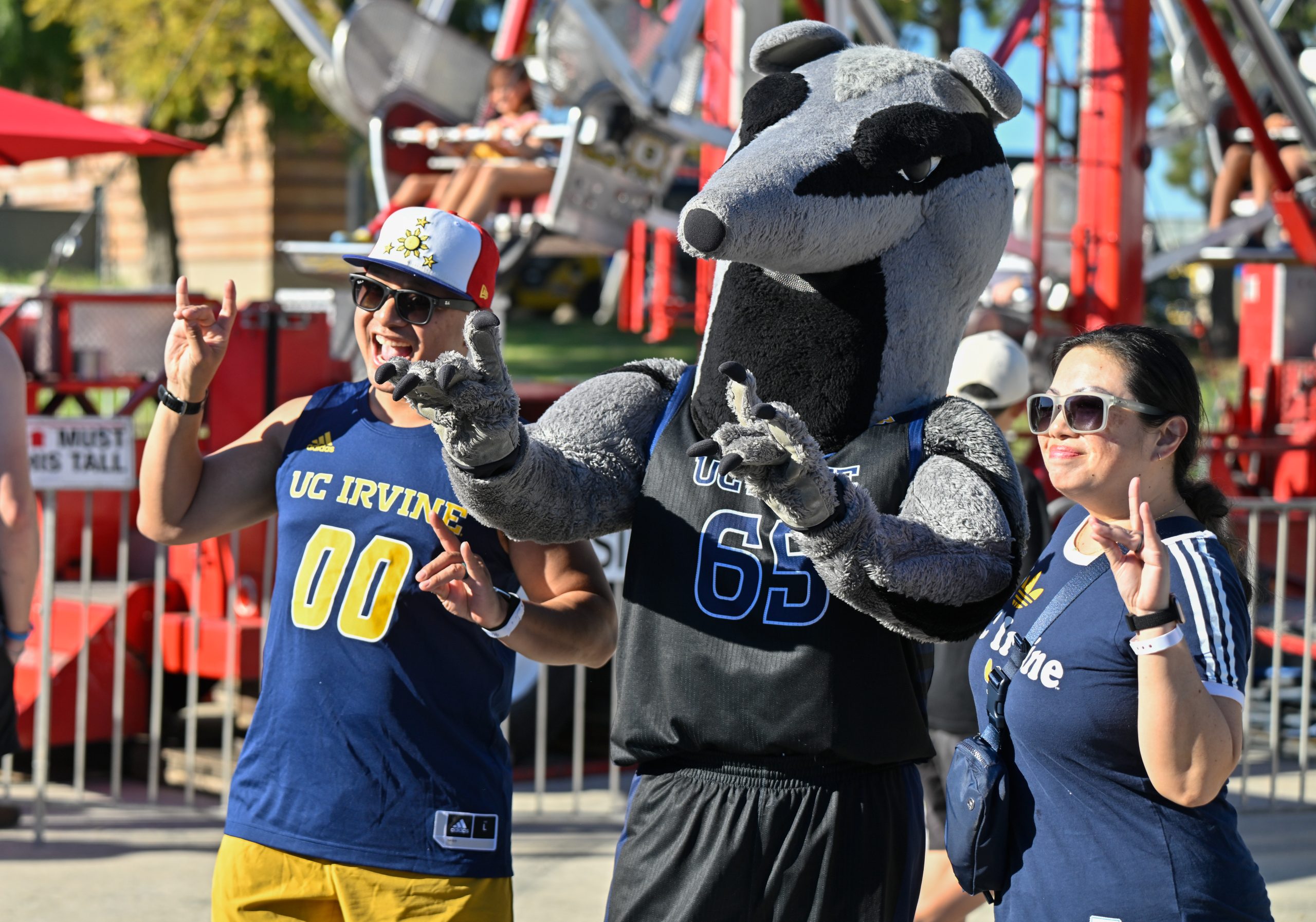 Three attendees pose with a UC Irvine anteater mascot in front of a carnival ride during a sunny outdoor homecoming celebration.