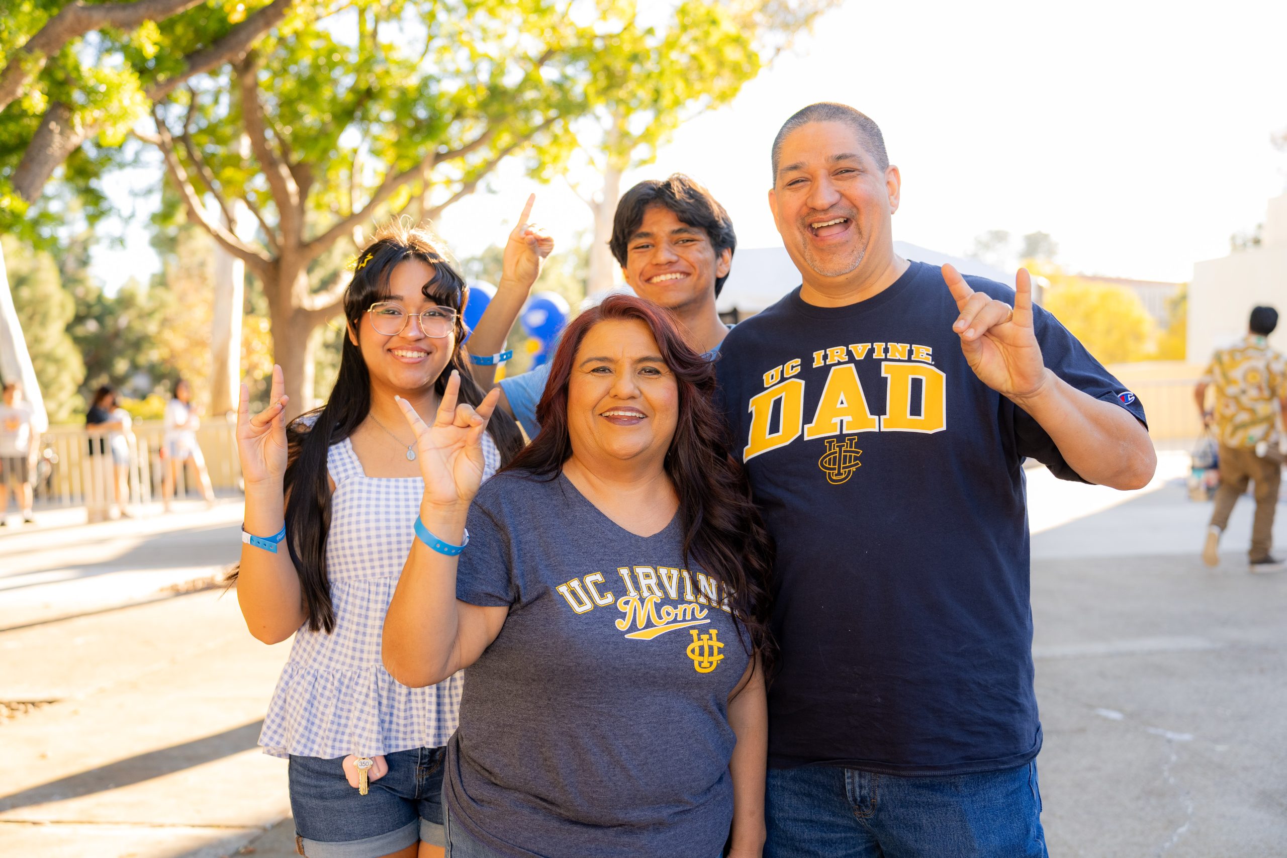 Four attendees at a UC Irvine homecoming event pose outdoors, making the UC Irvine hand sign while wearing UC Irvine-themed apparel.