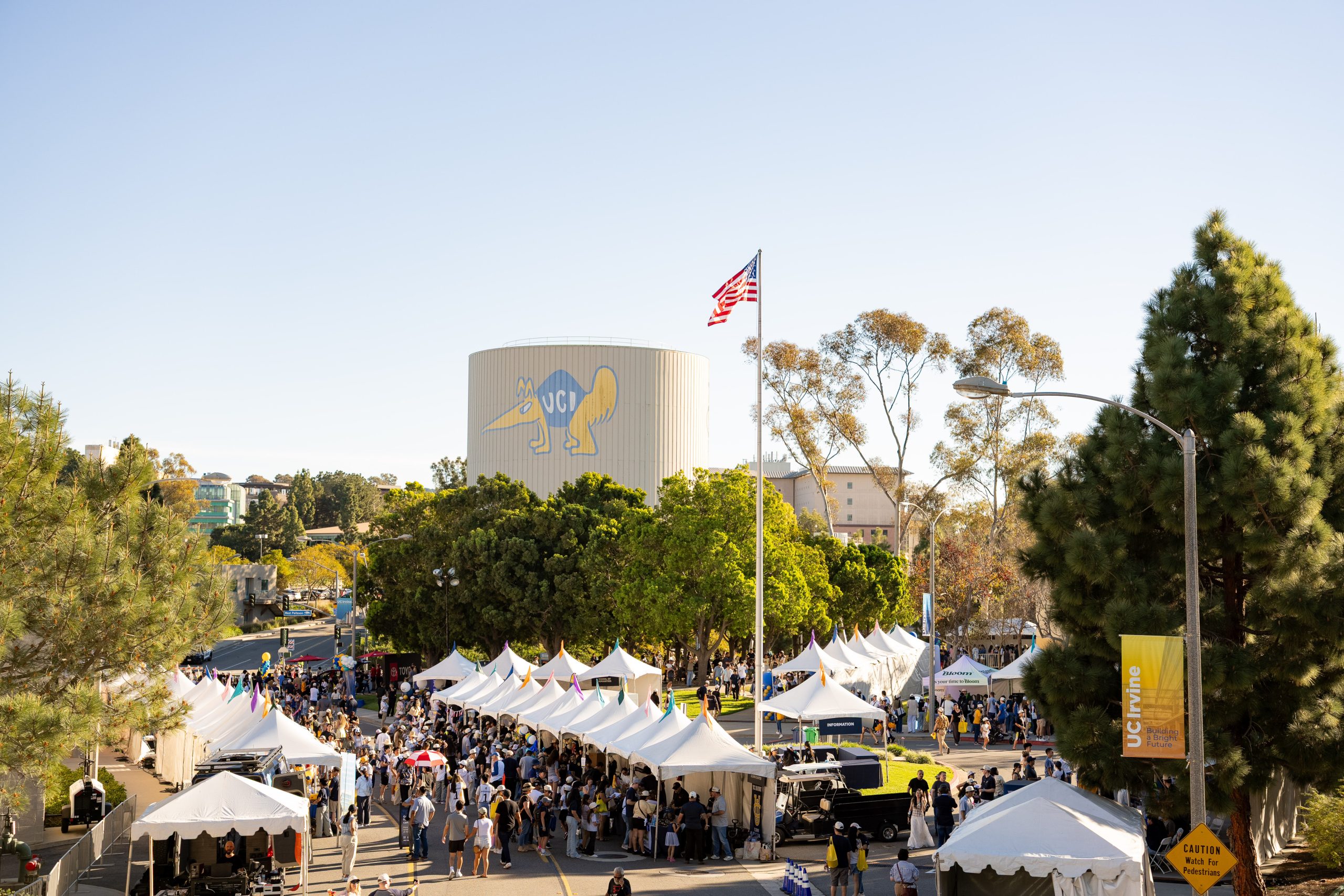 Large outdoor campus event with rows of white tents, hundreds of attendees, a UCI water tower, and an American flag rising above trees on a sunny day.