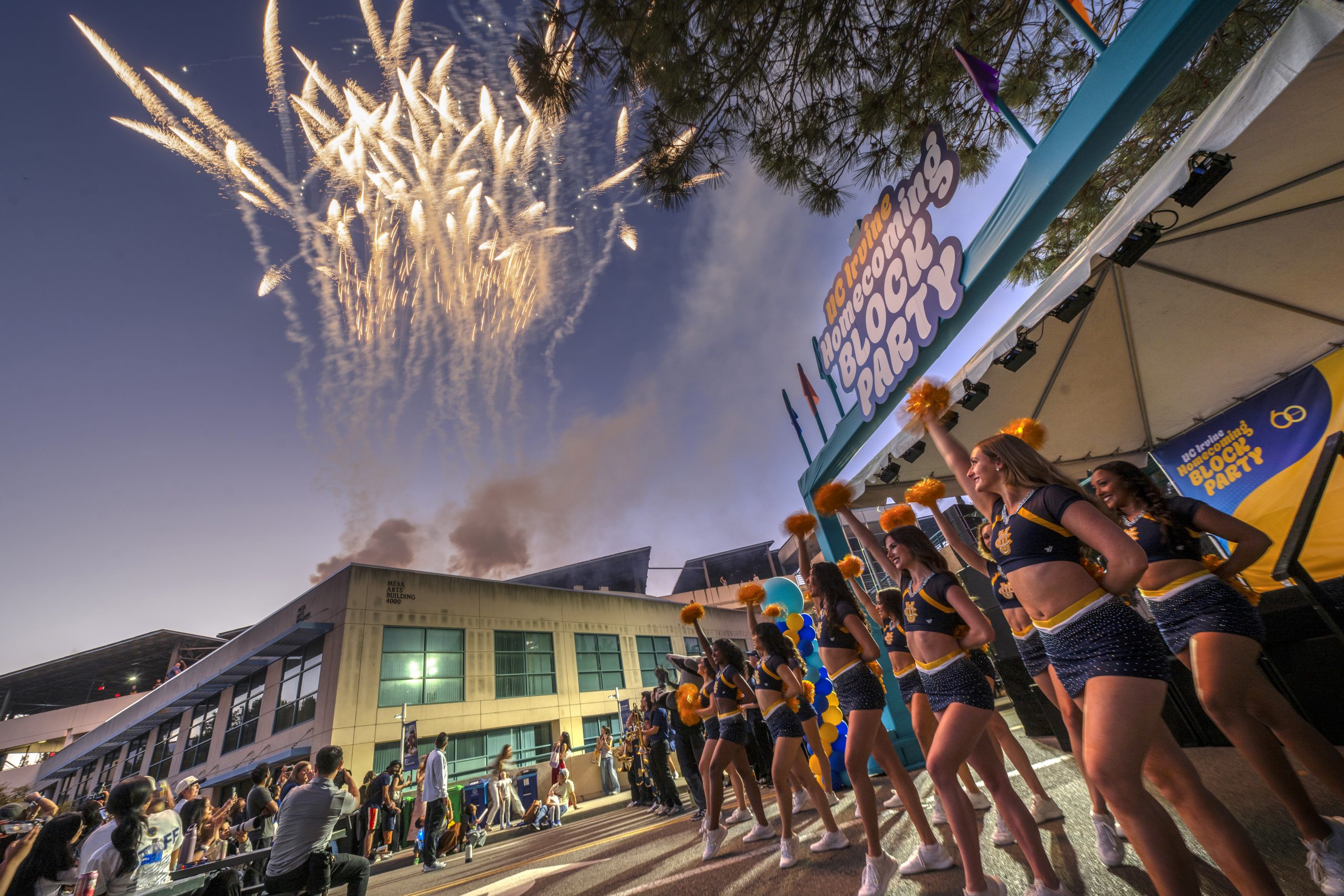 Fireworks light the evening sky above UC Irvine’s Homecoming Block Party as performers with gold pom-poms stand on an outdoor stage and spectators gather below.
