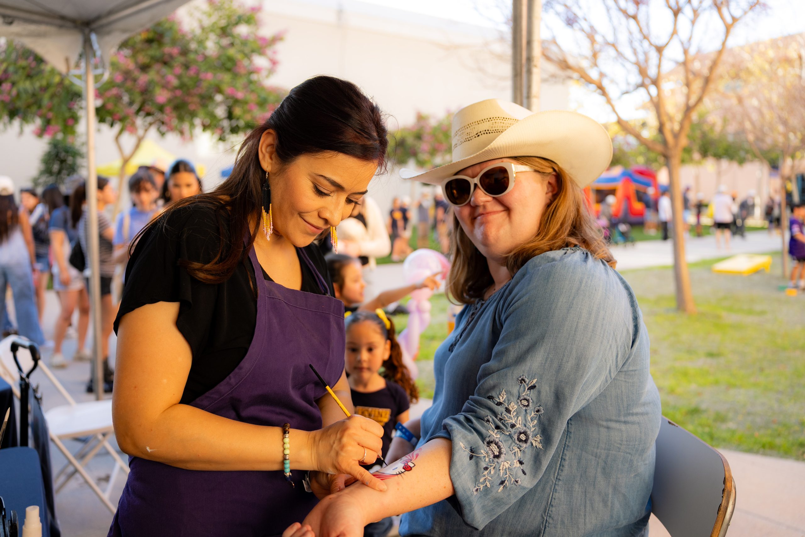 An attendee receives a colorful arm painting at an outdoor activity booth during a UC Irvine Homecoming event while families and visitors gather in the background.