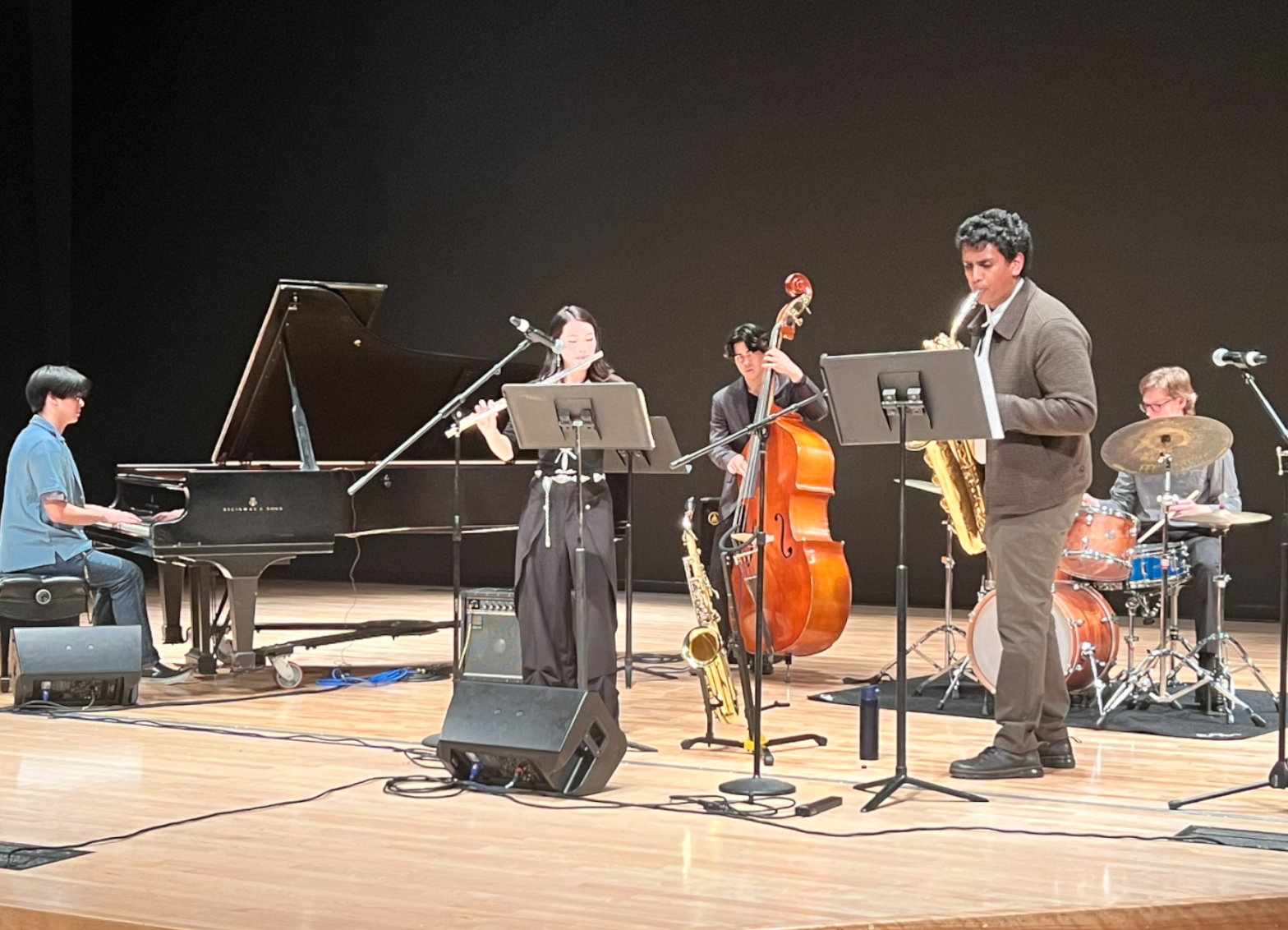 Small jazz ensemble performing on a concert hall stage, featuring piano, flute, upright bass, saxophone, and drums.