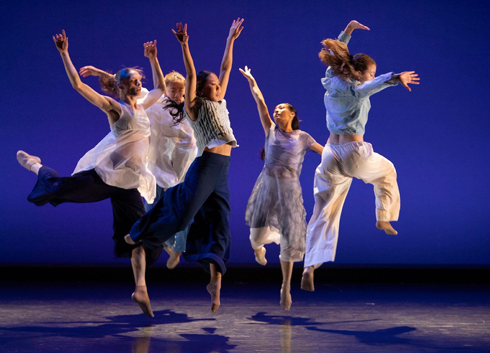 Dancers in flowing light-colored costumes leap across a stage under blue lighting, captured midair during a contemporary dance performance.