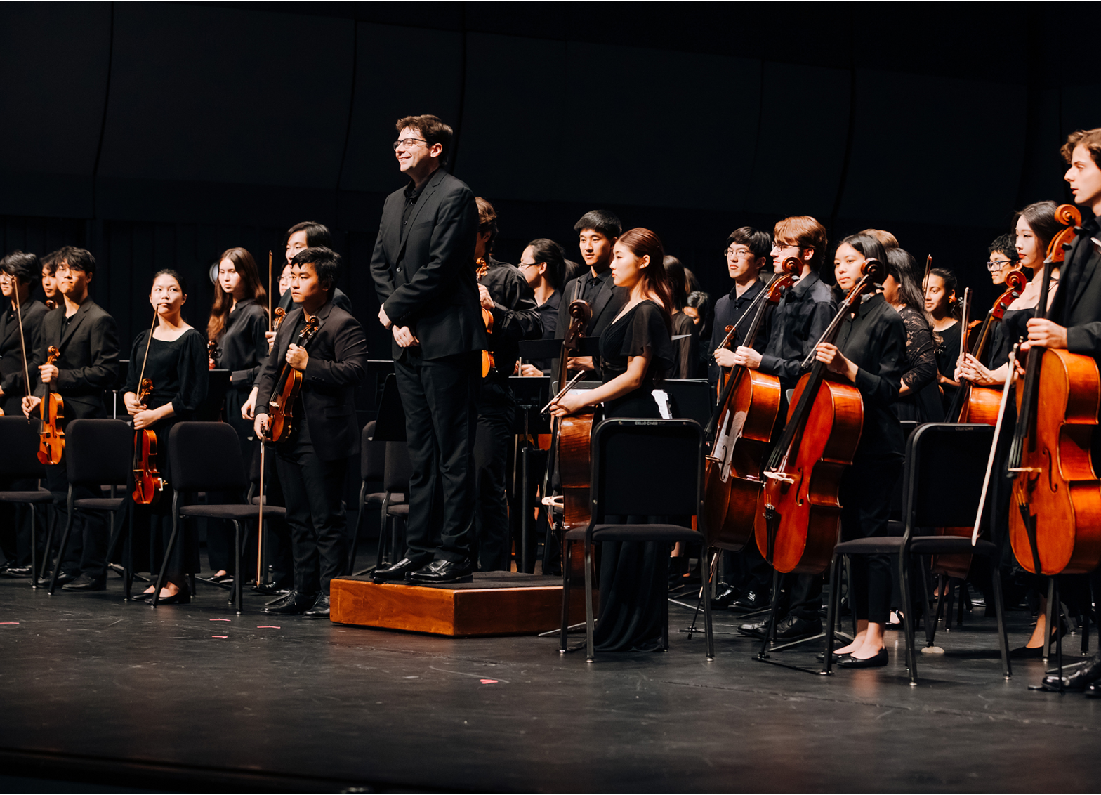 Student symphony orchestra stands onstage with string instruments as the conductor acknowledges the audience following a performance.