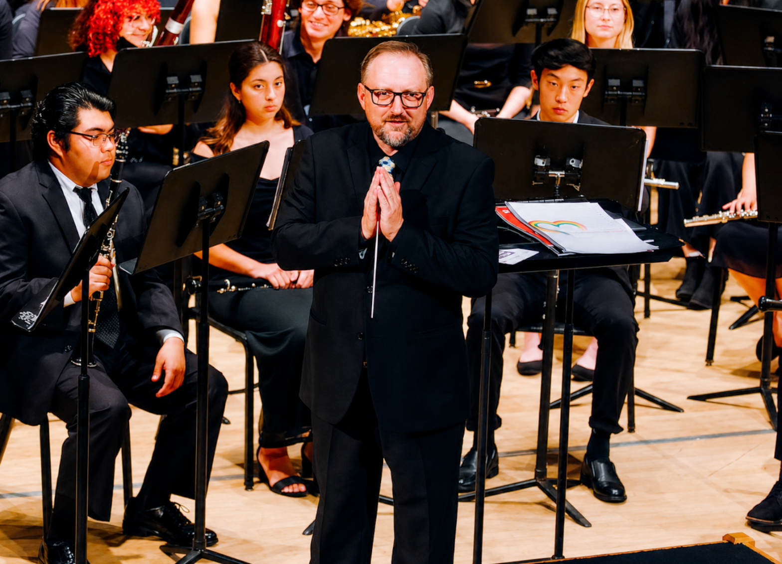 Conductor stands before a seated student wind ensemble on a concert hall stage, surrounded by musicians and music stands.