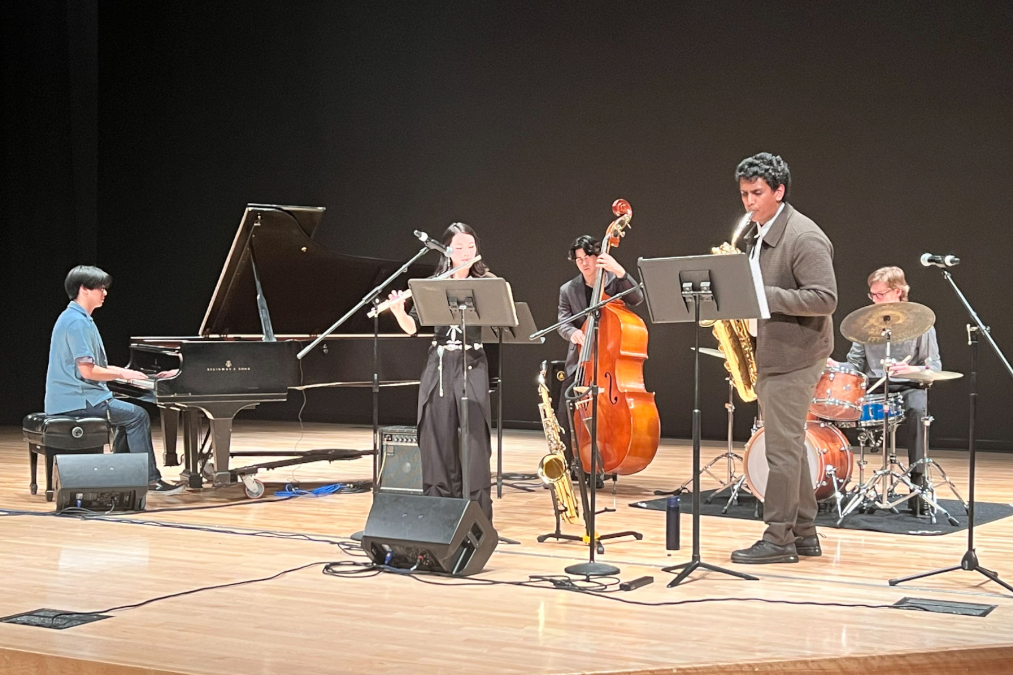 Small jazz ensemble performing on a concert hall stage, featuring piano, flute, upright bass, saxophone, and drums.
