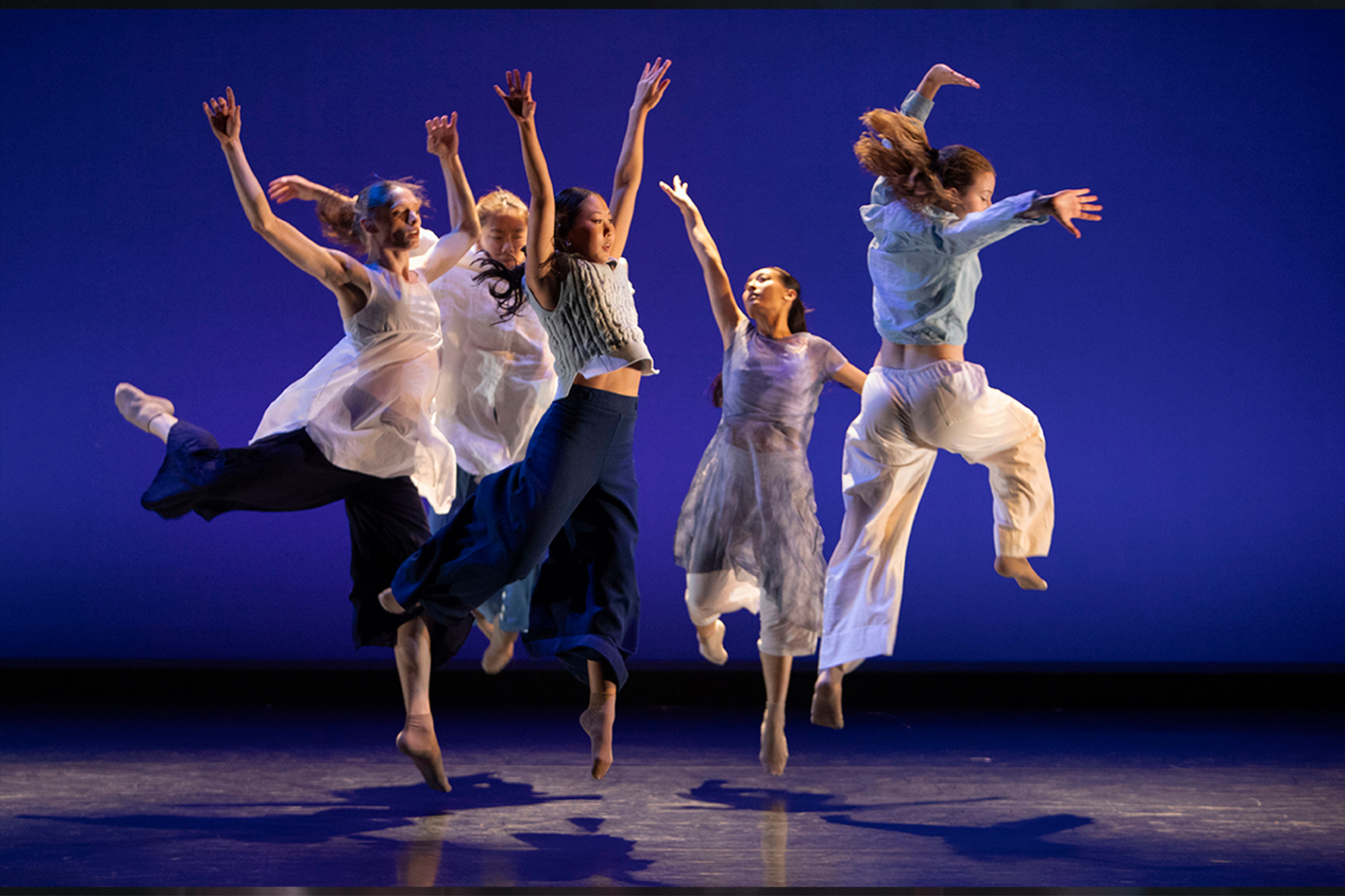Dancers in flowing light-colored costumes leap across a stage under blue lighting, captured midair during a contemporary dance performance.