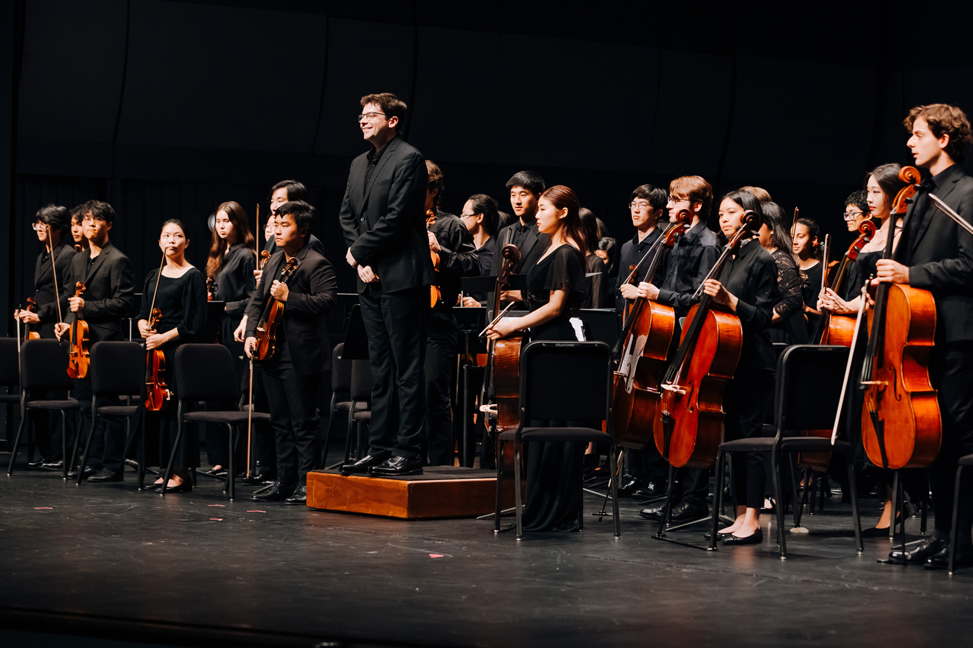 Student symphony orchestra stands onstage with string instruments as the conductor acknowledges the audience following a performance.