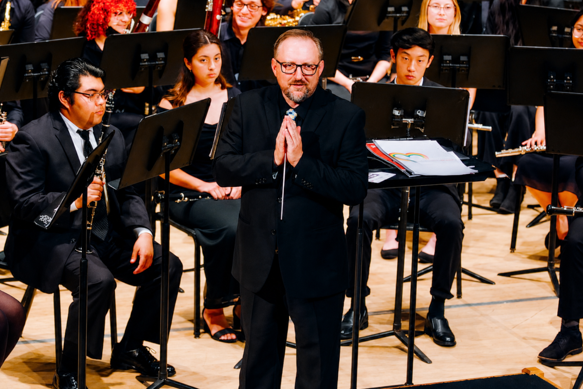 Conductor stands before a seated student wind ensemble on a concert hall stage, surrounded by musicians and music stands.